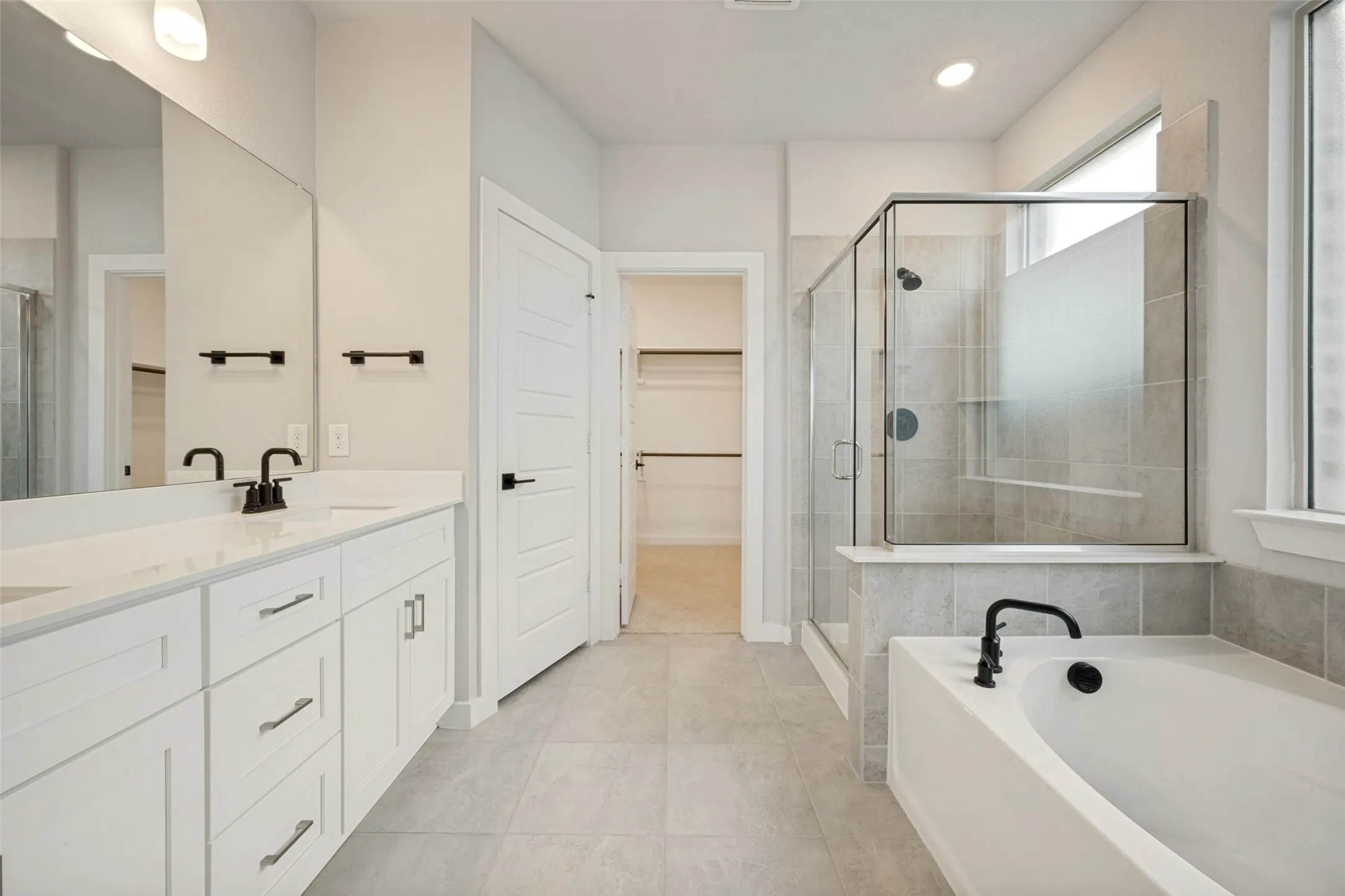 Bathroom featuring a dual vanity with white cabinetry and light-colored countertops, a separate walk-in shower with glass enclosure, a built-in soaking tub with matte black fixtures, and light grey tile flooring