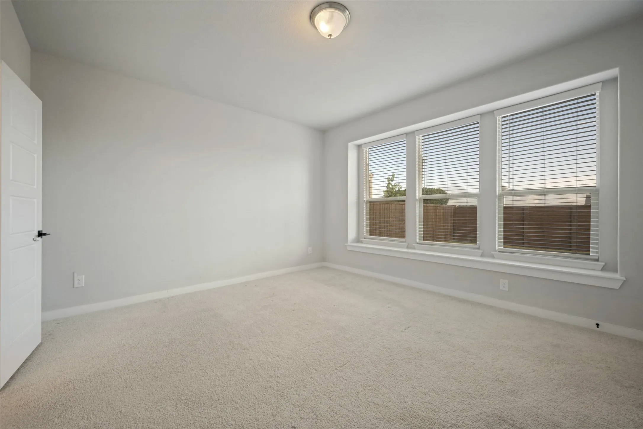 Spacious room featuring multiple large windows with horizontal blinds, neutral-toned carpet flooring, light gray wall paint, and a ceiling-mounted light fixture
