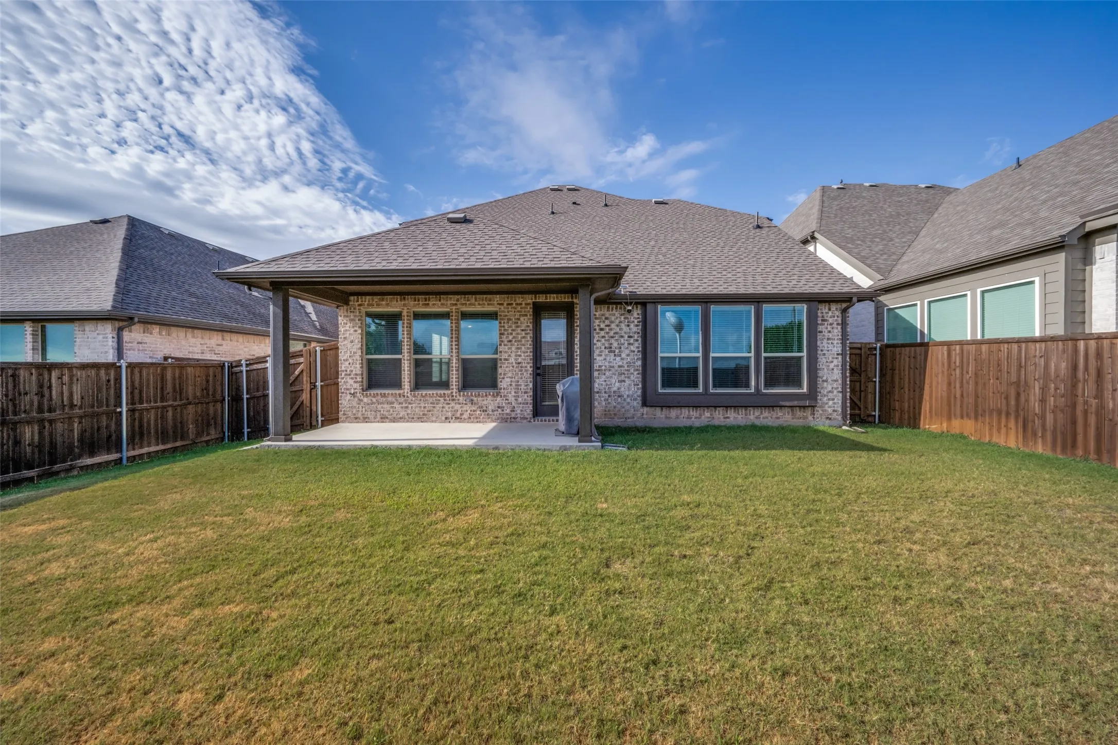 Expansive backyard featuring a manicured lawn, full perimeter privacy fencing, covered patio with concrete flooring, multiple windows with dark trim, and a brick exterior