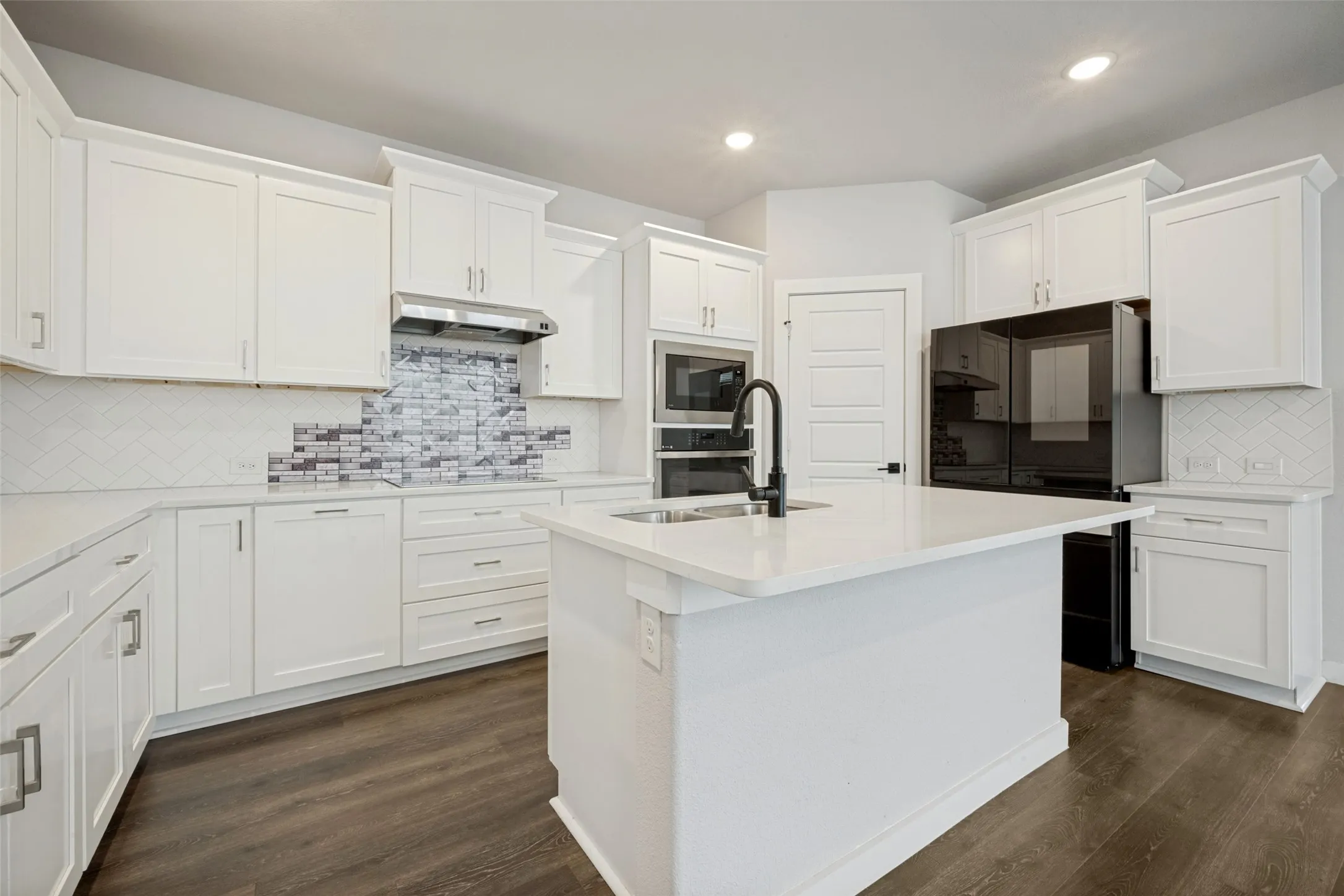 Kitchen featuring a central island with an integrated sink and matte black faucet, surrounded by white cabinetry with contrasting hardware, light countertops, and a subway tile backsplash