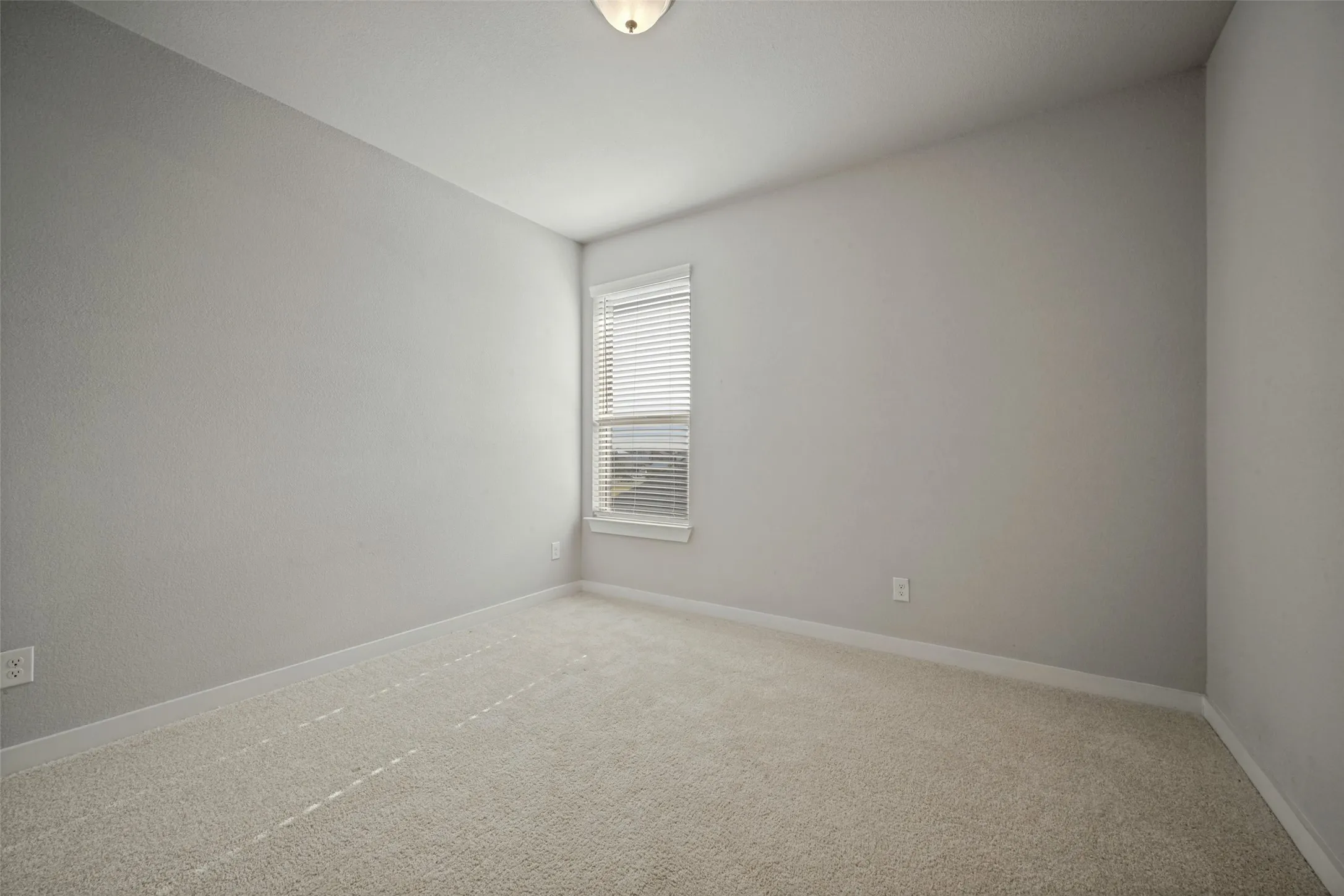 Neutral-toned room featuring a single window with horizontal blinds, light-colored carpeting, a flush-mount ceiling light fixture, and white baseboards