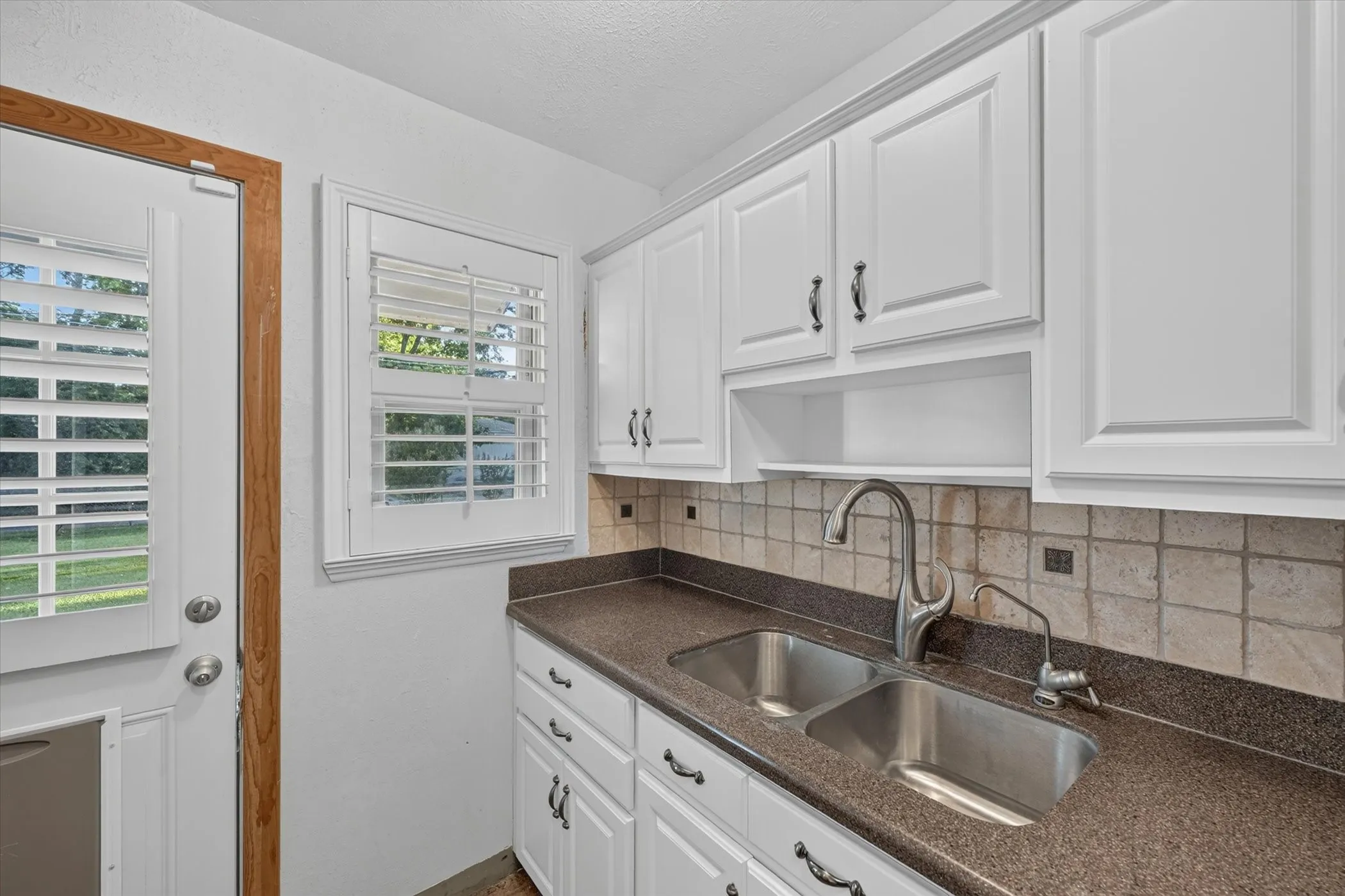 Kitchen featuring white shaker-style cabinetry, a double basin stainless steel sink with a gooseneck faucet, a light-toned tile backsplash, and dark stone-finish countertops
