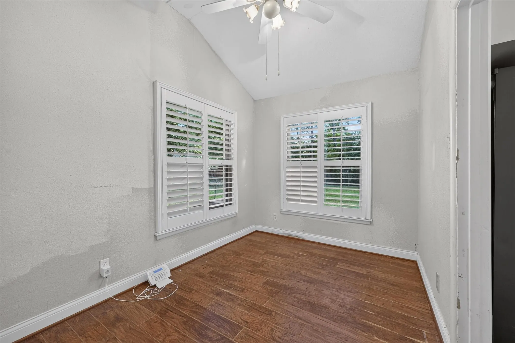 Room featuring wood-finish flooring, two windows with plantation shutters, a vaulted ceiling, and a ceiling fan with light fixture