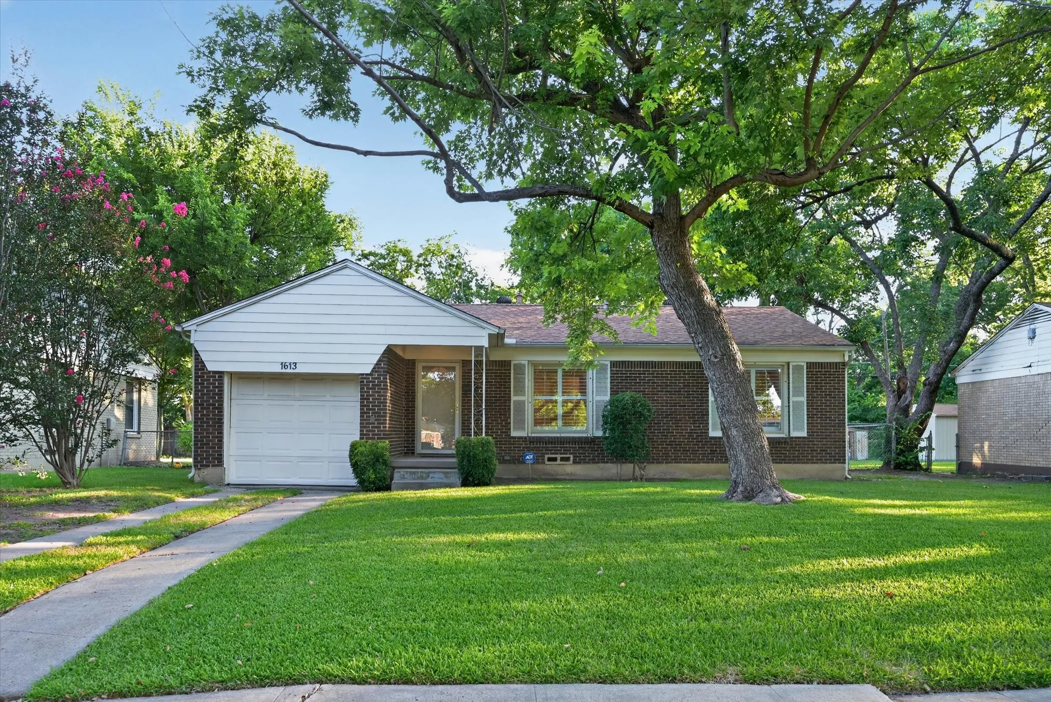 Brick facade residence featuring a front-facing attached garage, covered entryway, mature front yard tree, manicured lawn, and concrete driveway
