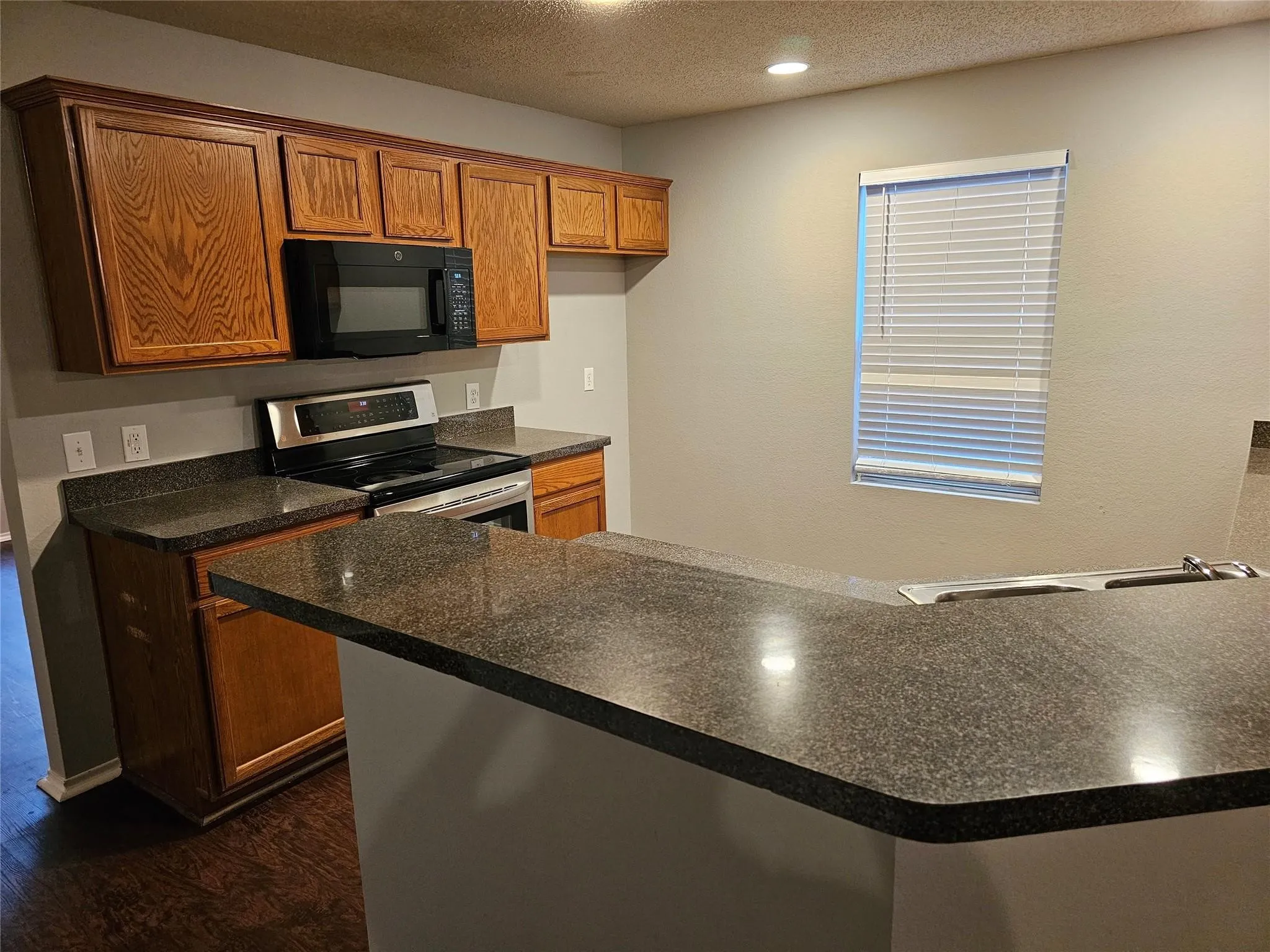 Kitchen with brown cabinets, a textured ceiling, stainless steel range with electric cooktop, black microwave, and dark countertops
