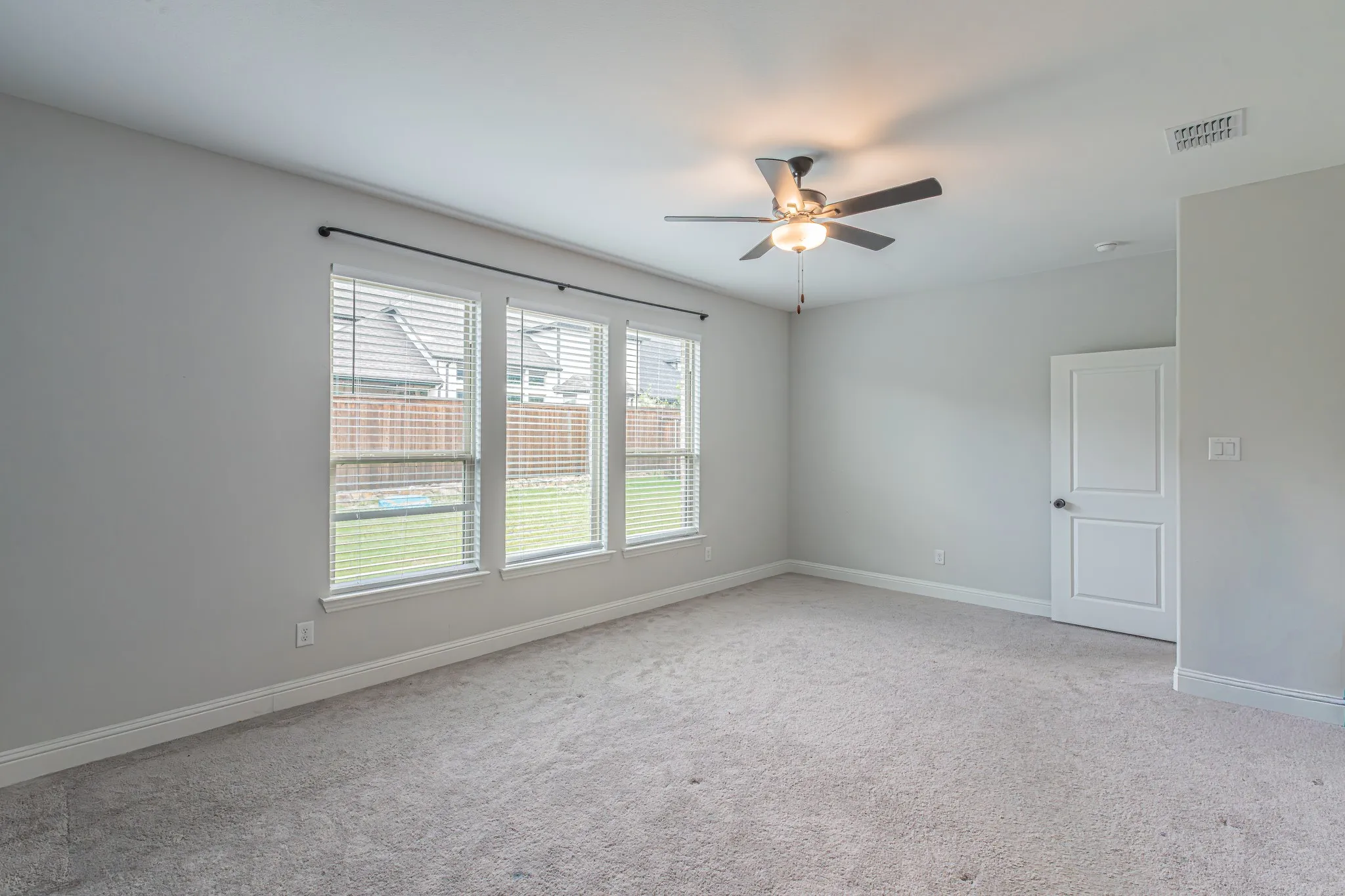 Carpeted interior space featuring a ceiling fan with integrated lighting, three windows with horizontal blinds, and a white paneled door