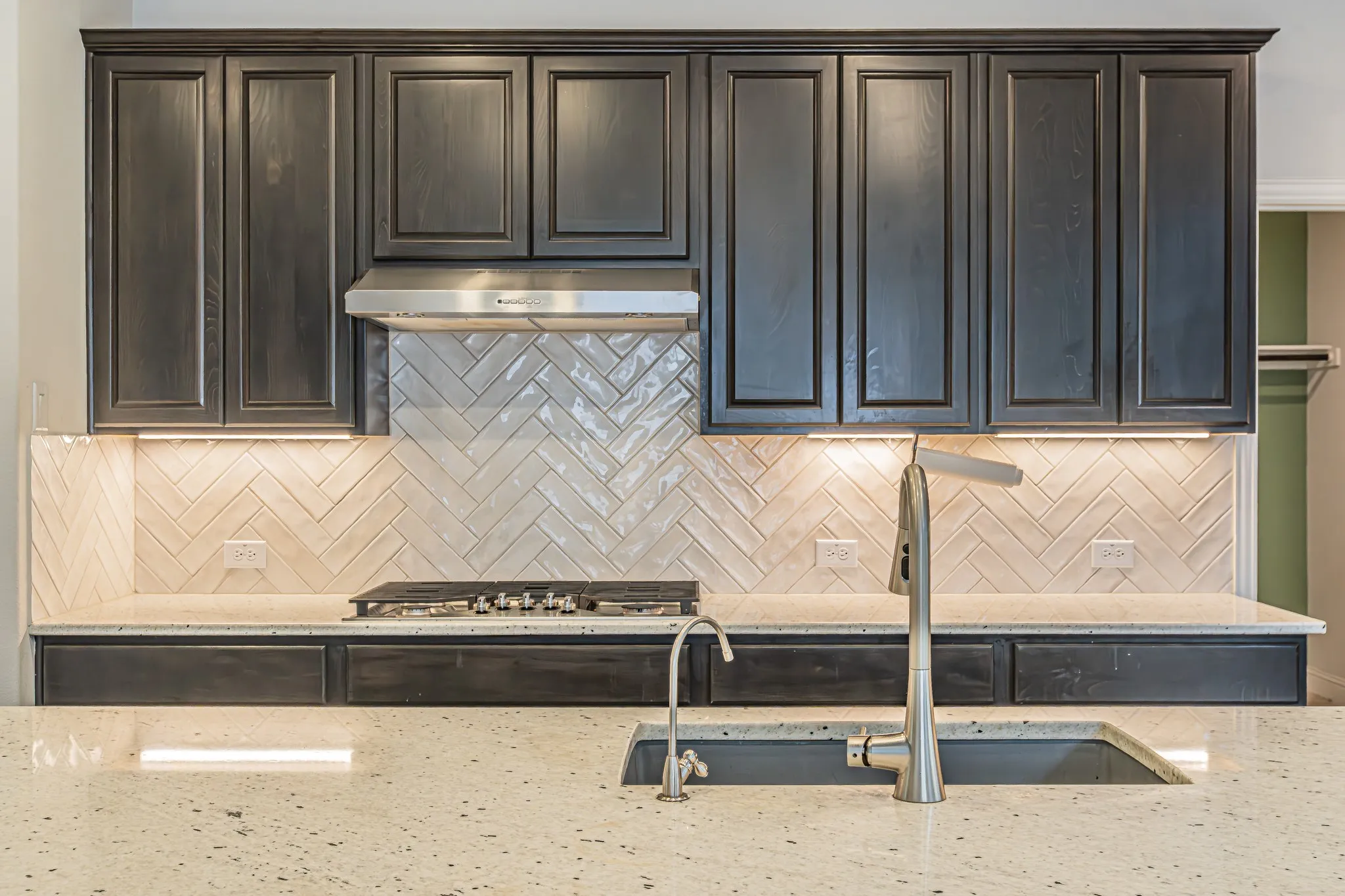 Kitchen featuring dark wood cabinetry, a herringbone pattern tile backsplash, under-cabinet lighting, a built-in gas range, and a stainless steel range hood