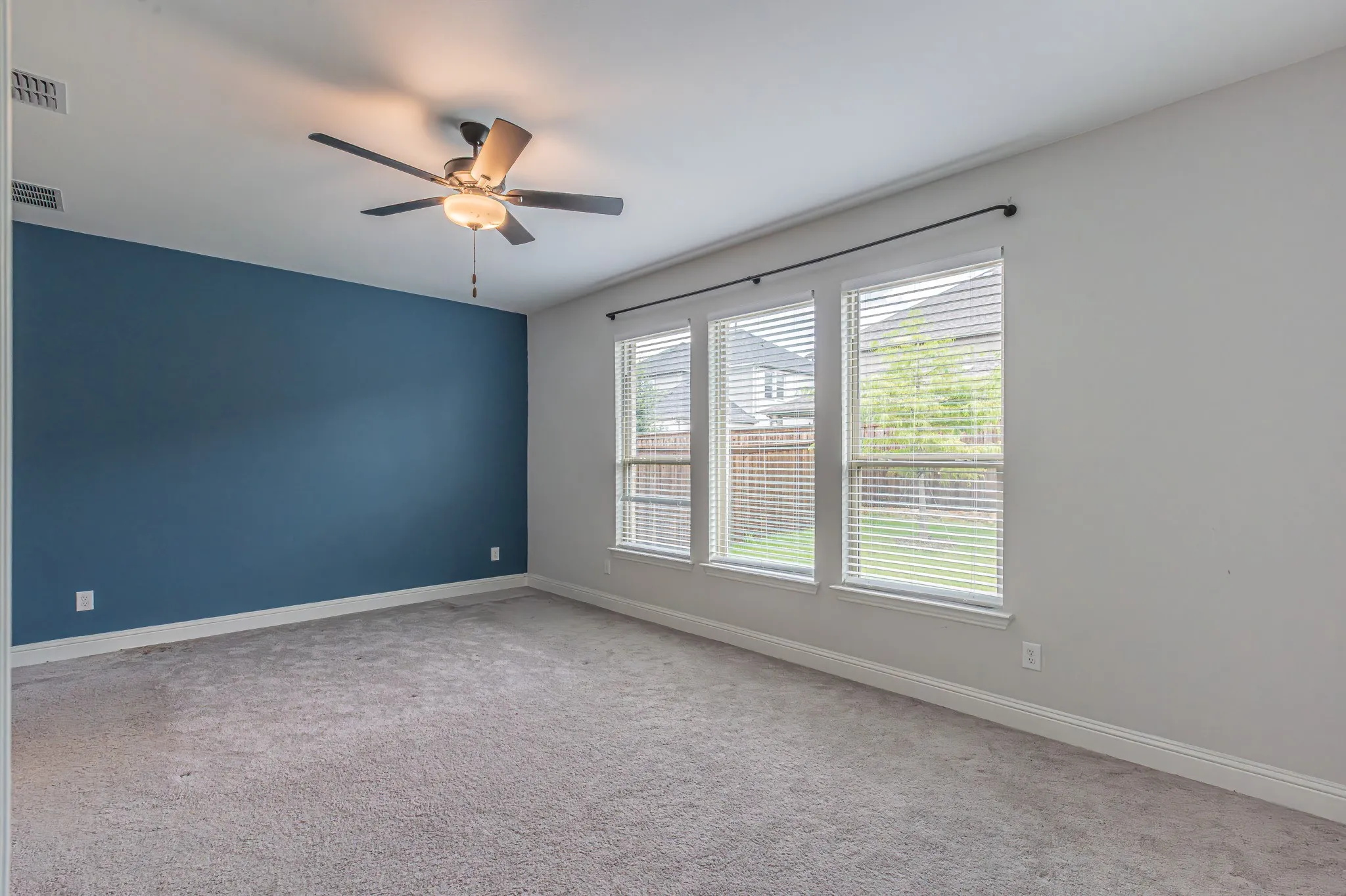Spacious room featuring a blue accent wall, three large windows with blinds, light-toned carpeting, and a ceiling fan with integrated lighting
