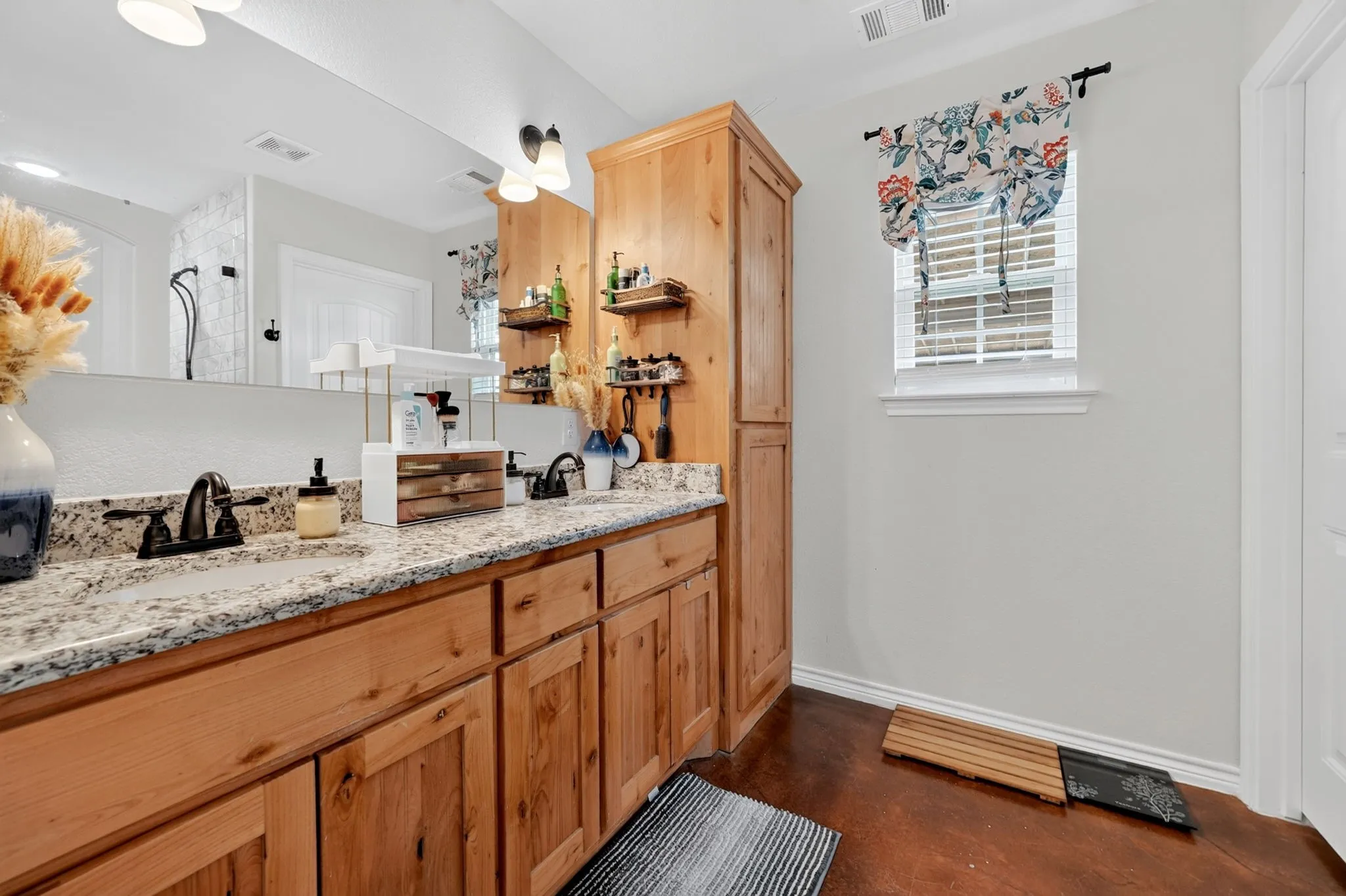 Dual vanity featuring light-toned wood cabinetry with multiple storage options, granite countertops, and dark bronze fixtures