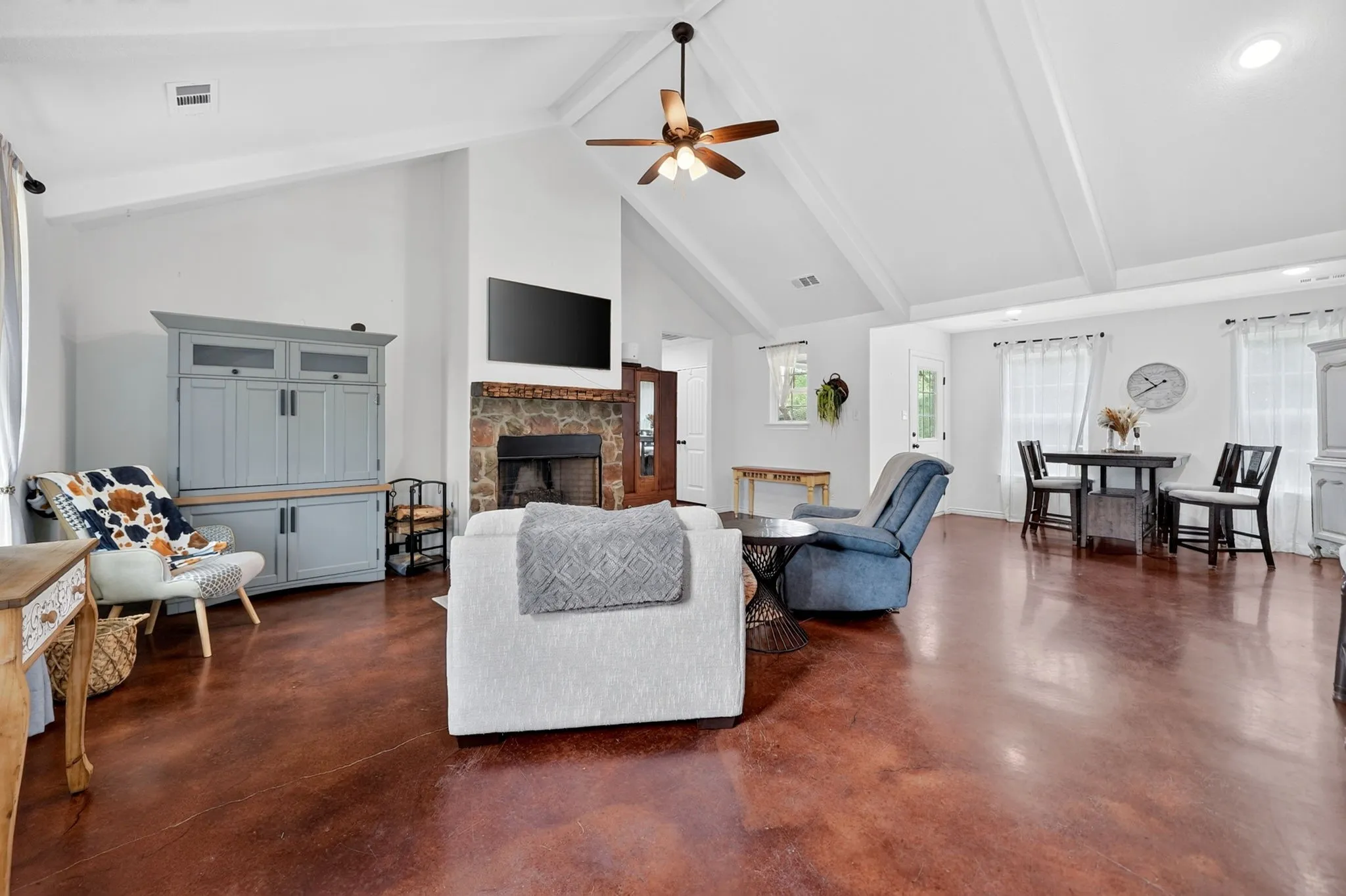 Open-concept living area featuring vaulted ceilings with exposed beams, stained concrete flooring, a stone-faced fireplace, and a ceiling fan
