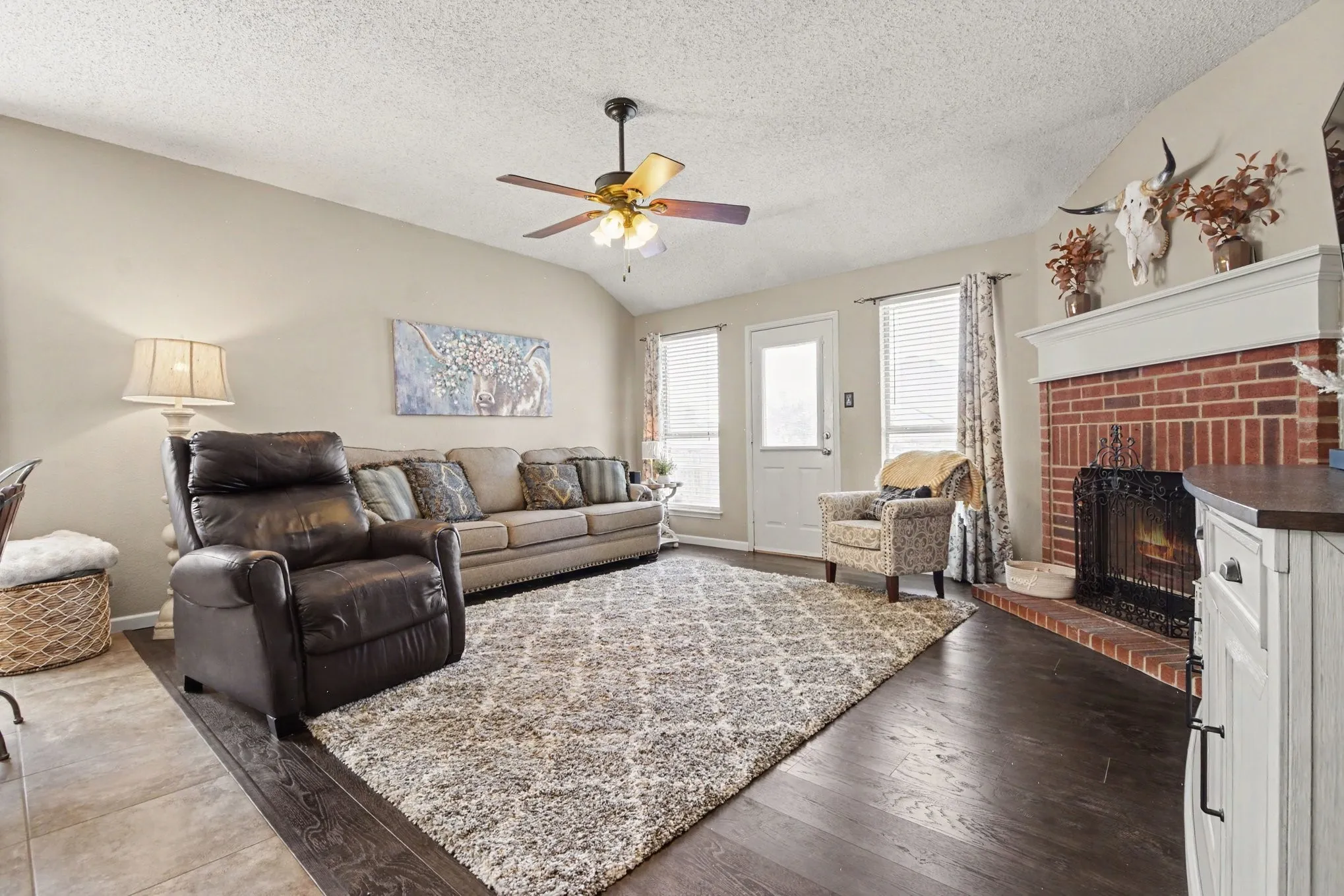 Spacious living area featuring high vaulted ceilings, rich wood-finish flooring, and a brick fireplace with a white mantel