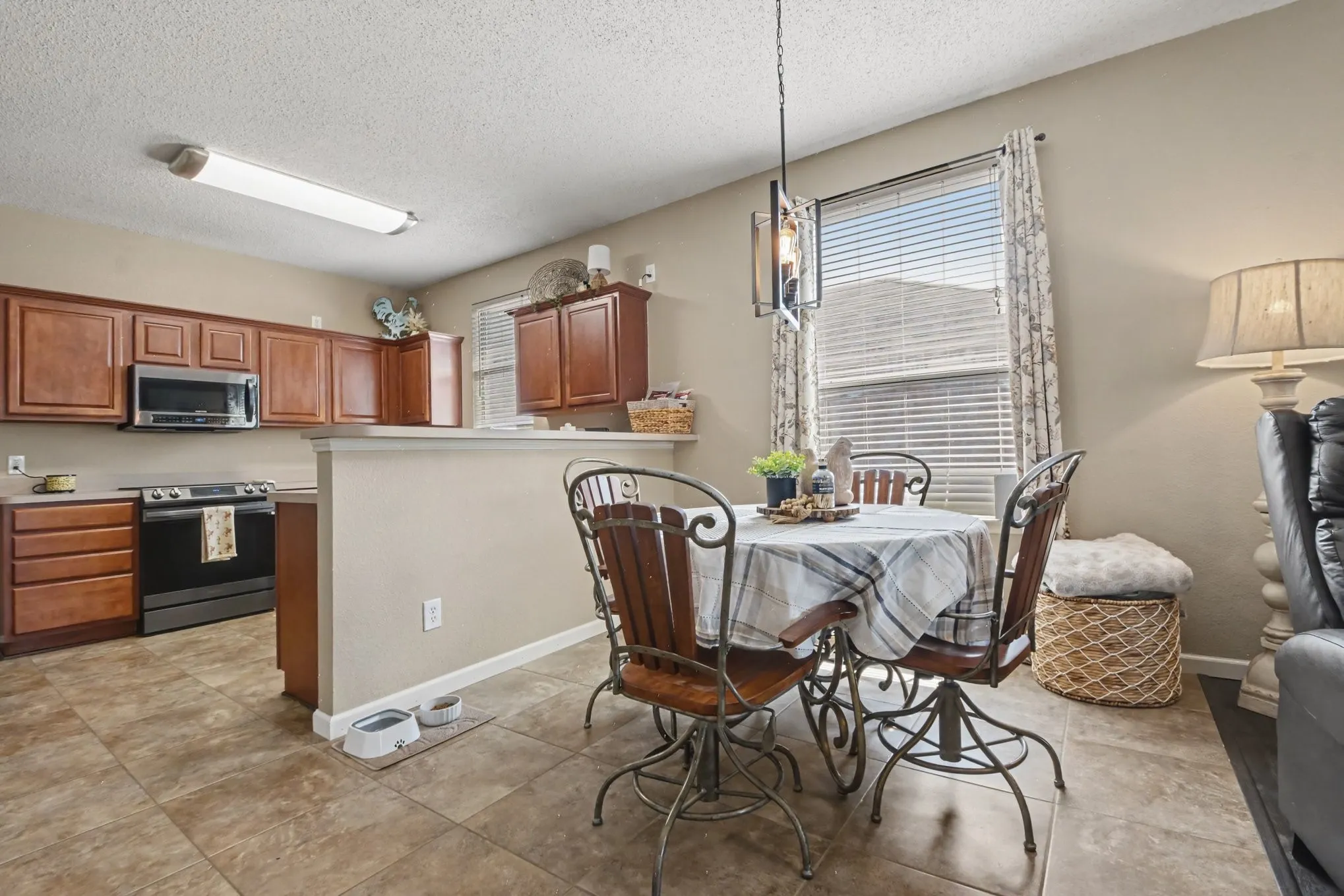 Open-concept kitchen and dining area featuring warm wood cabinetry, stainless steel appliances, a breakfast bar, tile flooring, and a dining area with a modern chandelier and window with blinds