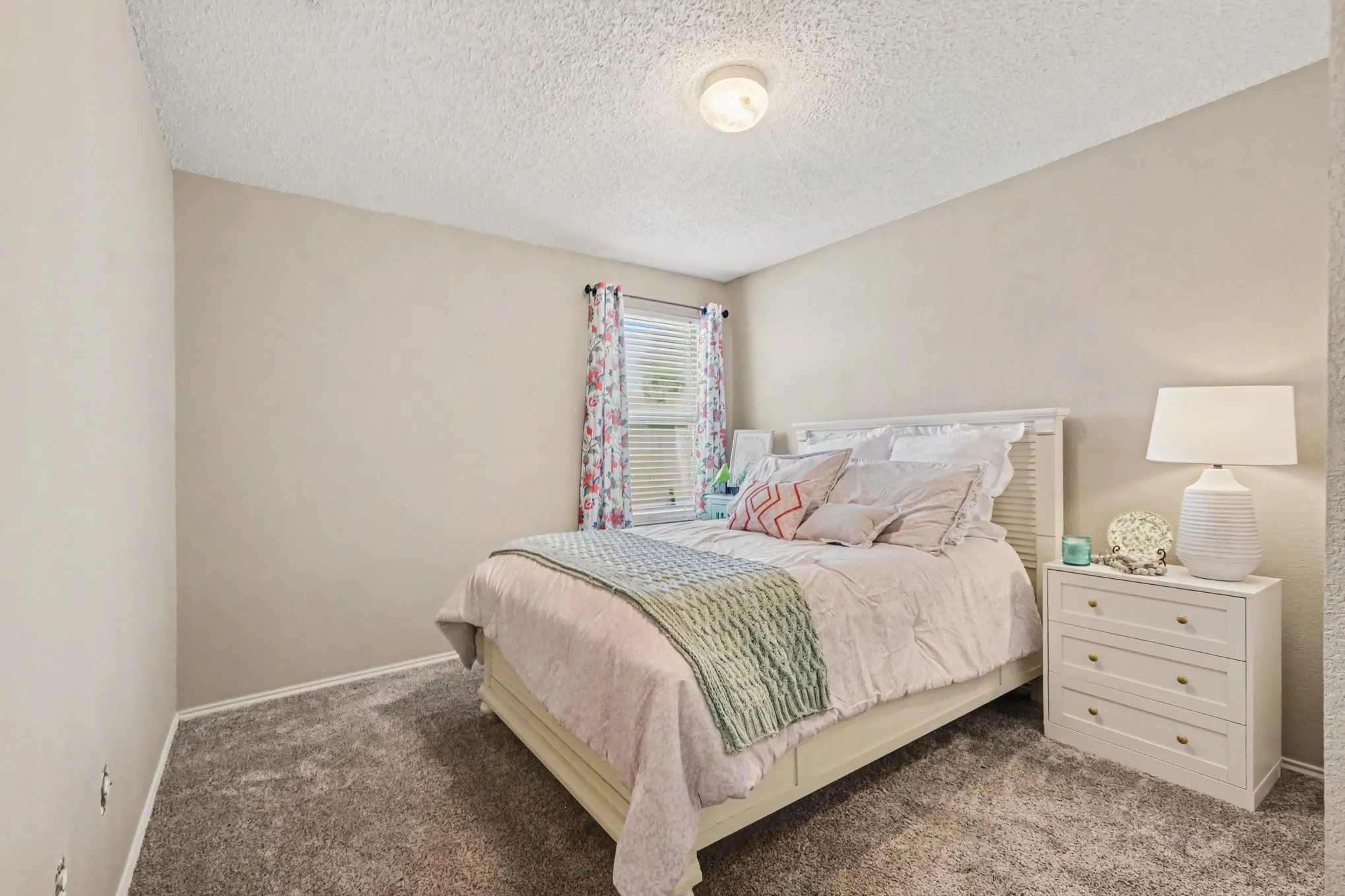 Neutral wall paint throughout, plush gray carpeting, and a window with blinds provide a comfortable setting