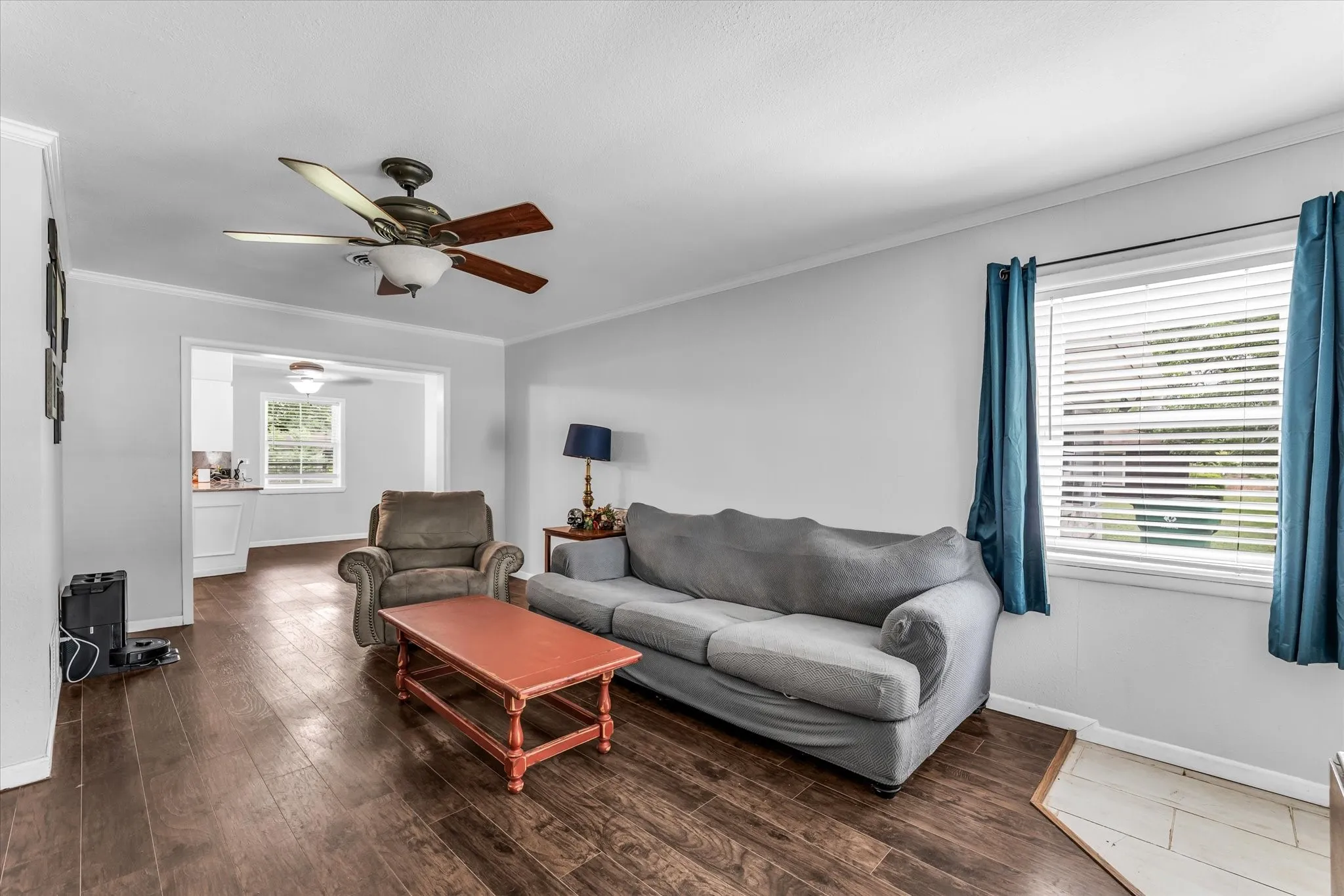 Spacious living area featuring wood-finish flooring, a ceiling fan with integrated lighting, and a broad window with horizontal blinds