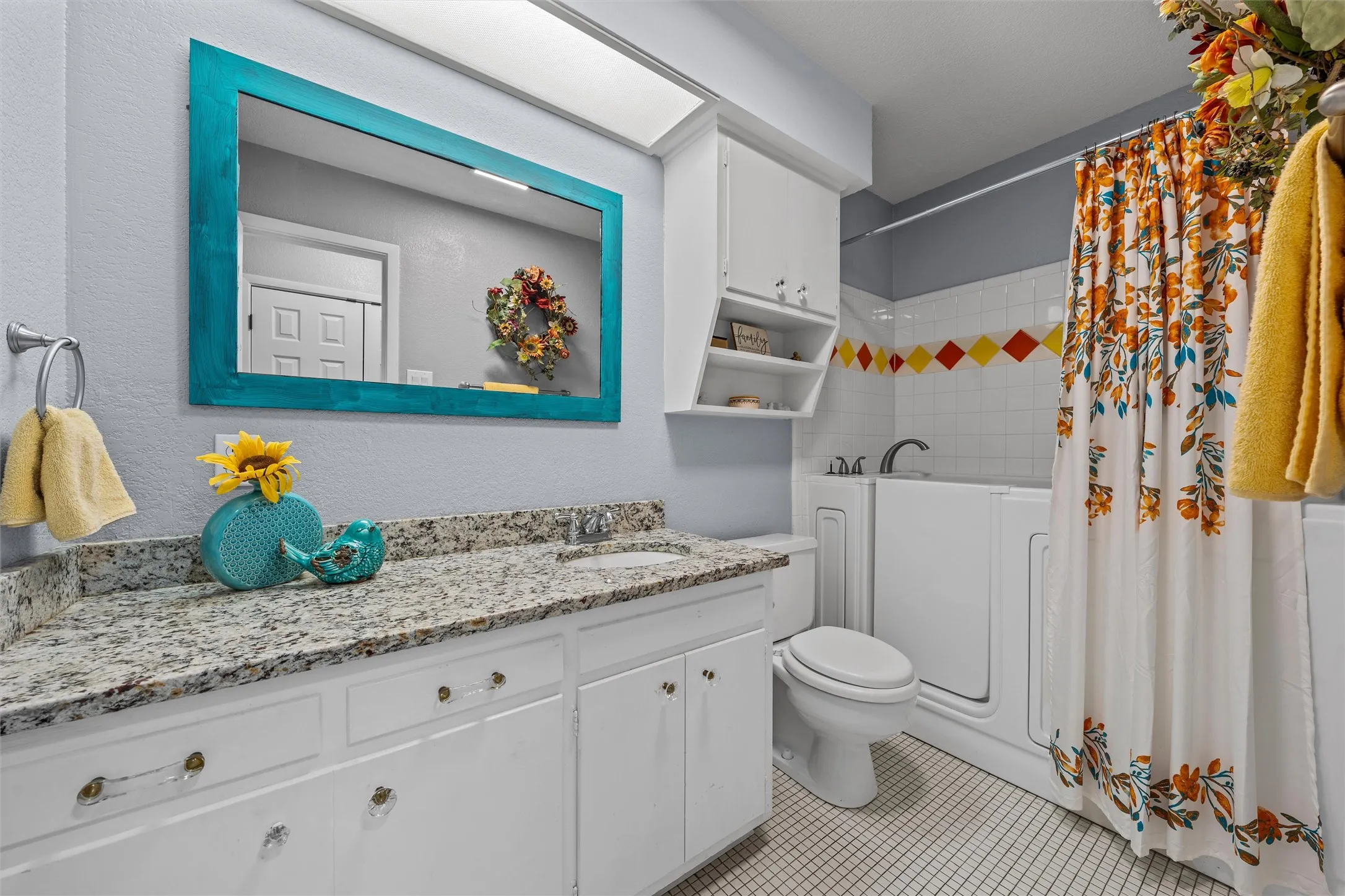 Bathroom featuring a white vanity with a granite-style countertop, a rectangular turquoise-framed mirror, a walk-in bathtub, white wall tiles with decorative accents, and white grid-patterned floor tiles