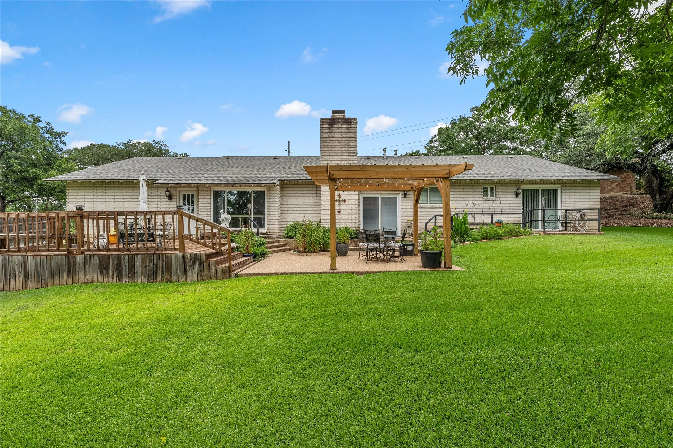 Expansive backyard featuring a large wood-finish deck, a ground-level patio with a pergola, a brick exterior, a substantial chimney, and mature trees