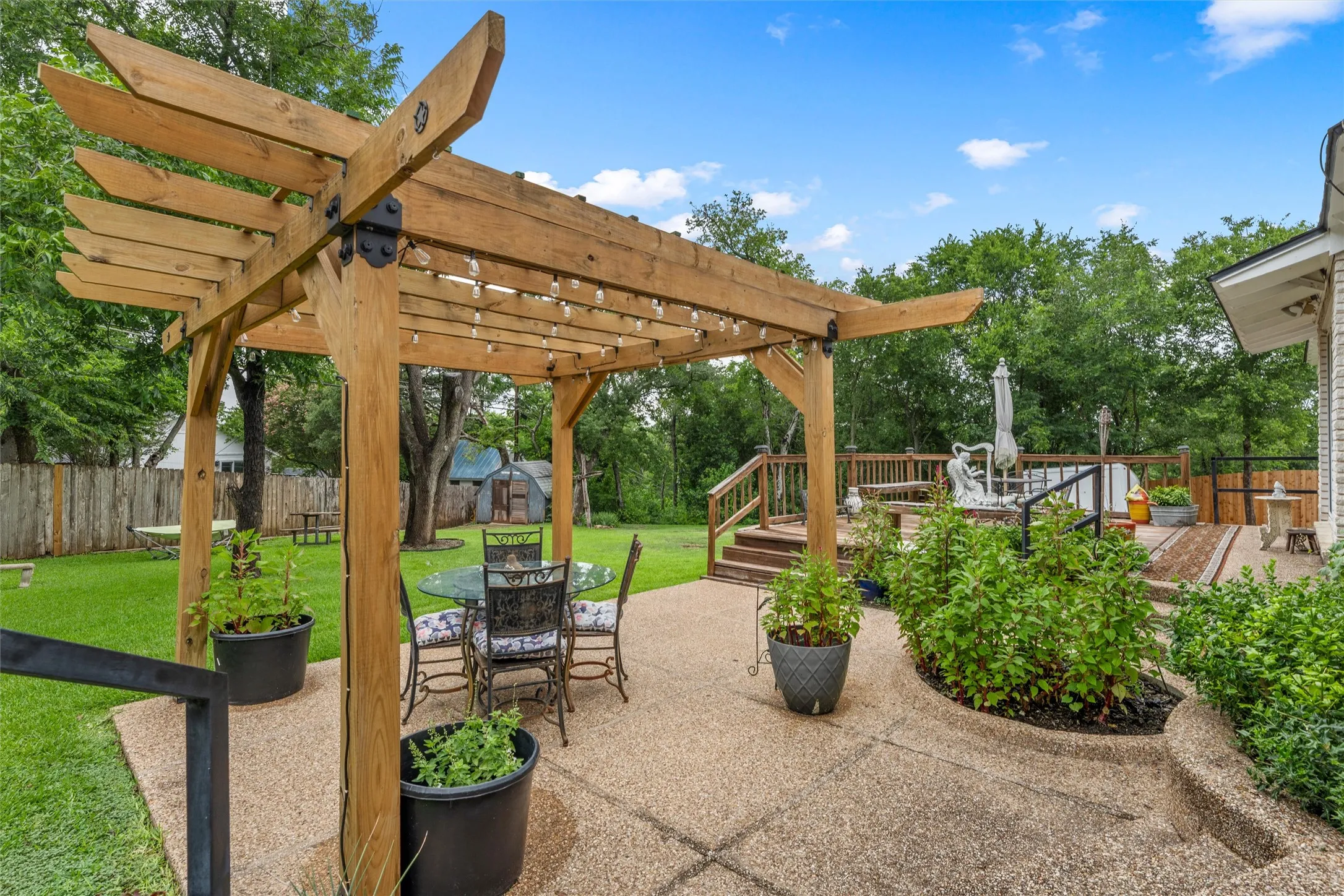 Expansive outdoor living area featuring a wooden pergola, an aggregate patio, a tiered wooden deck, mature landscaping, and a substantial fenced yard