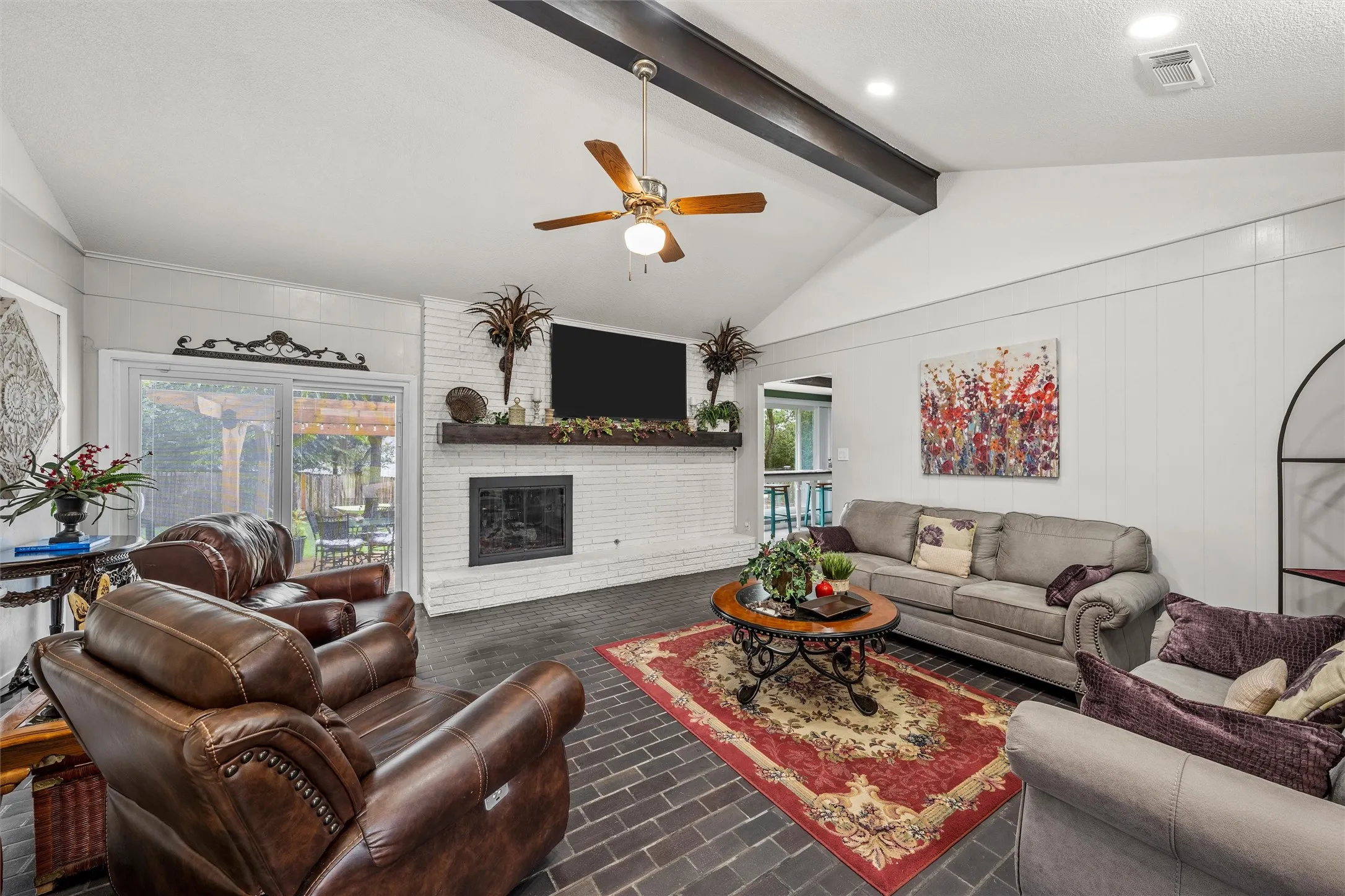Vaulted ceiling living area featuring dark wood-finish beams, a white painted brick fireplace with a wood mantel, and dark brick flooring