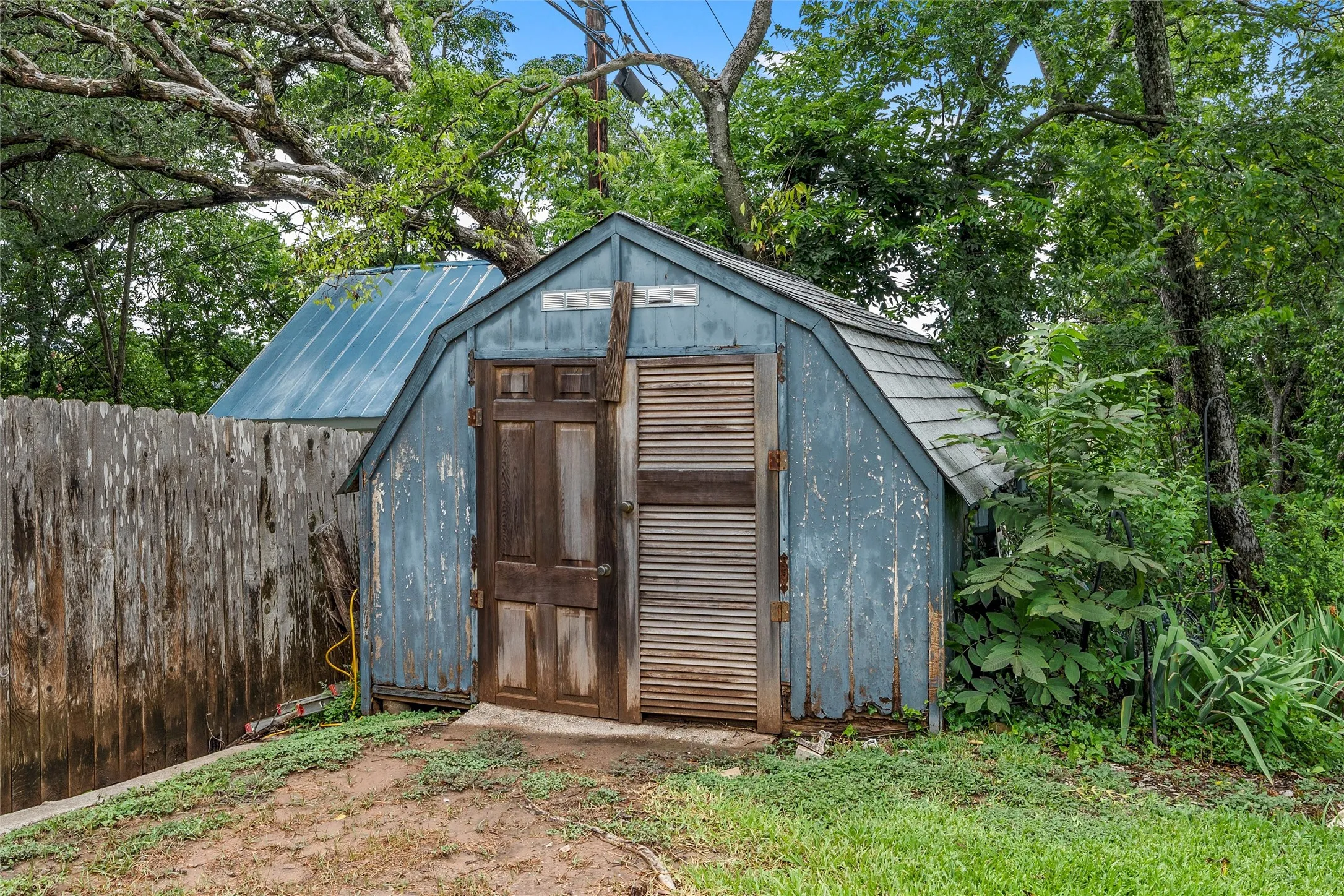 Detached storage shed with a gambrel-style roof, single wood panel door, and louvered panel door, situated on a grassy area with mature trees and a wood fence