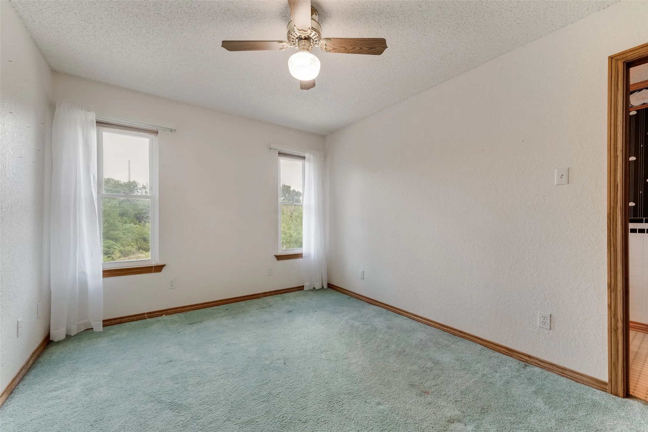Room featuring two windows with white curtains, a ceiling fan with integrated lighting, textured white walls, and a light blue carpet