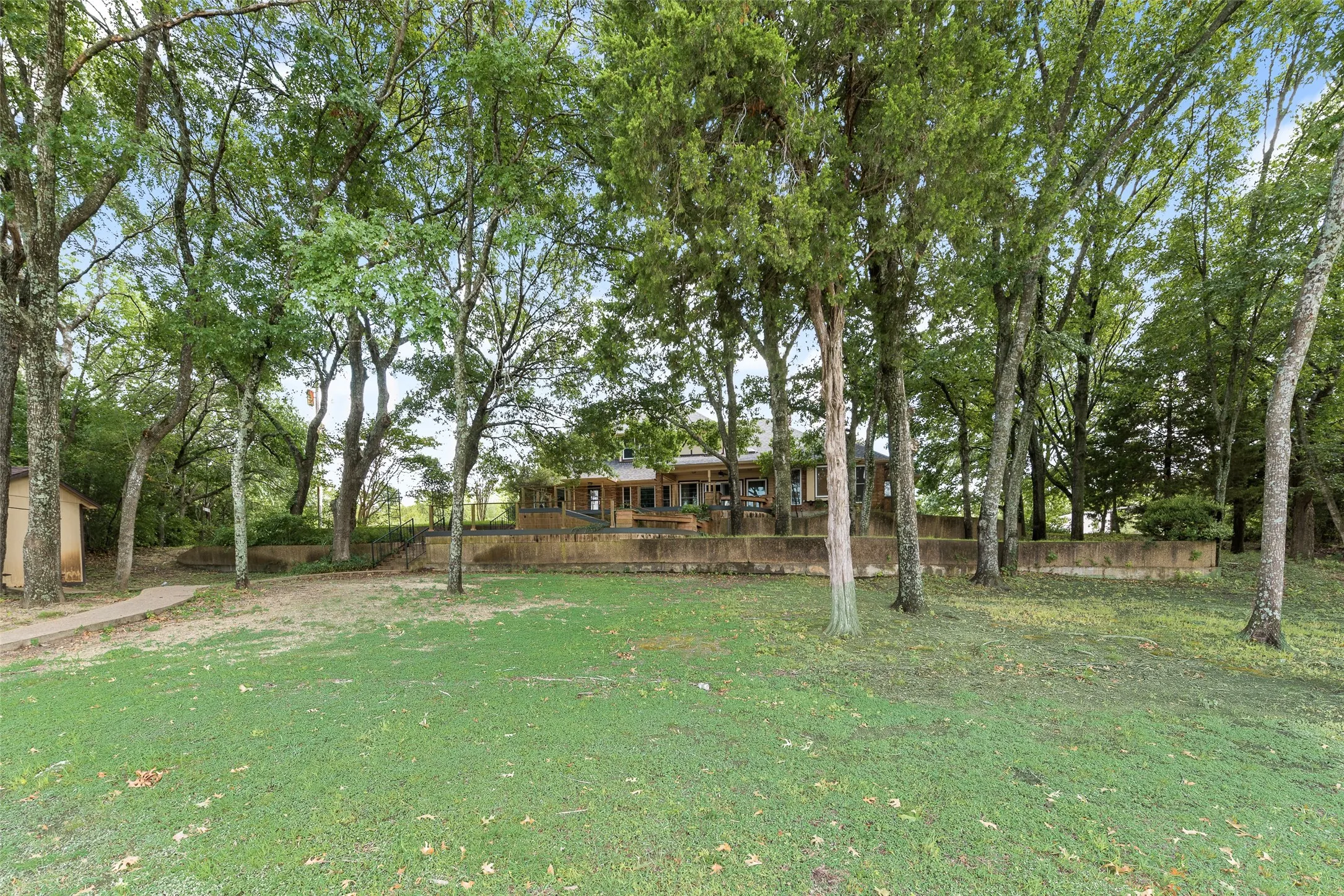 Expansive outdoor area featuring a lush green lawn, mature trees providing natural shade, a stone pathway, and a detached shed