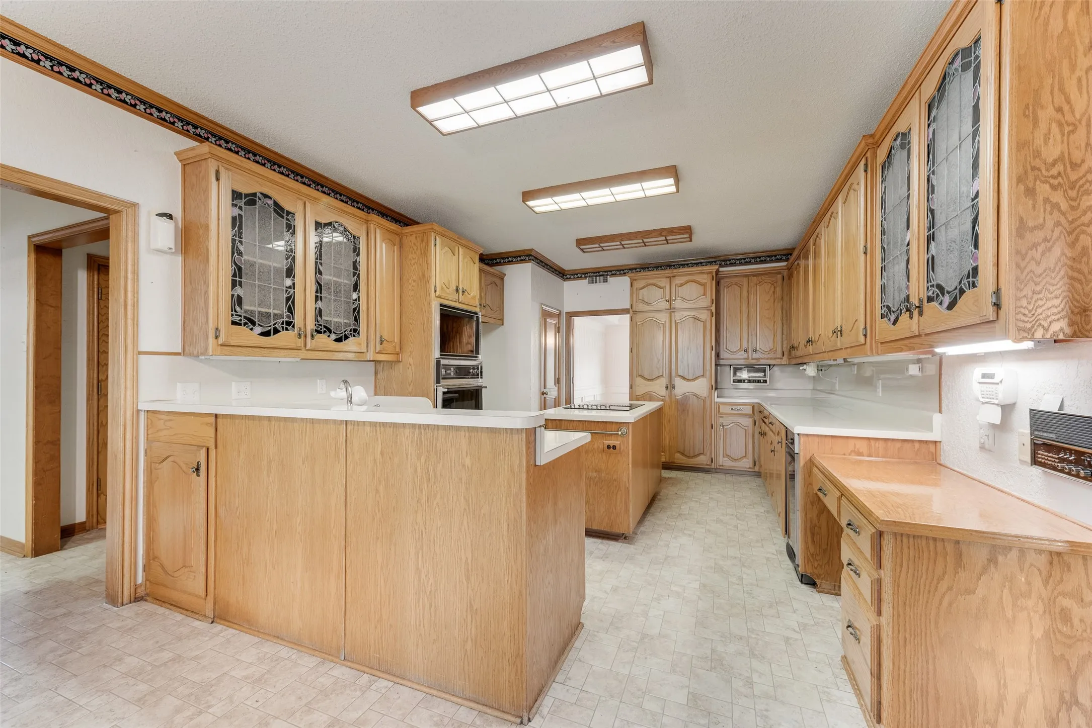 Spacious kitchen featuring abundant wood cabinetry, white countertops, built-in appliances, a central island, and patterned resilient flooring