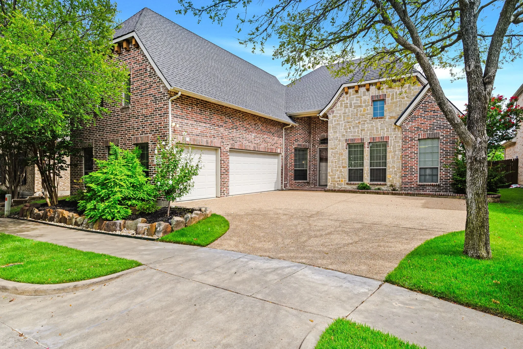 Brick and stone facade residence featuring a two-car garage, mature landscaping, and a concrete driveway