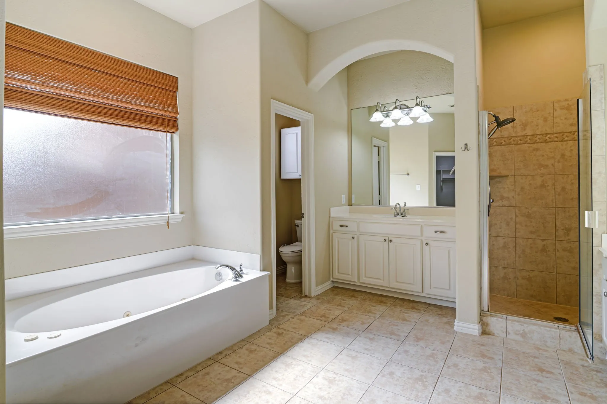 Spacious bathroom featuring a built-in soaking tub with a frosted privacy window, a tiled walk-in shower with a glass enclosure, and a vanity with dual sinks under an arched mirror
