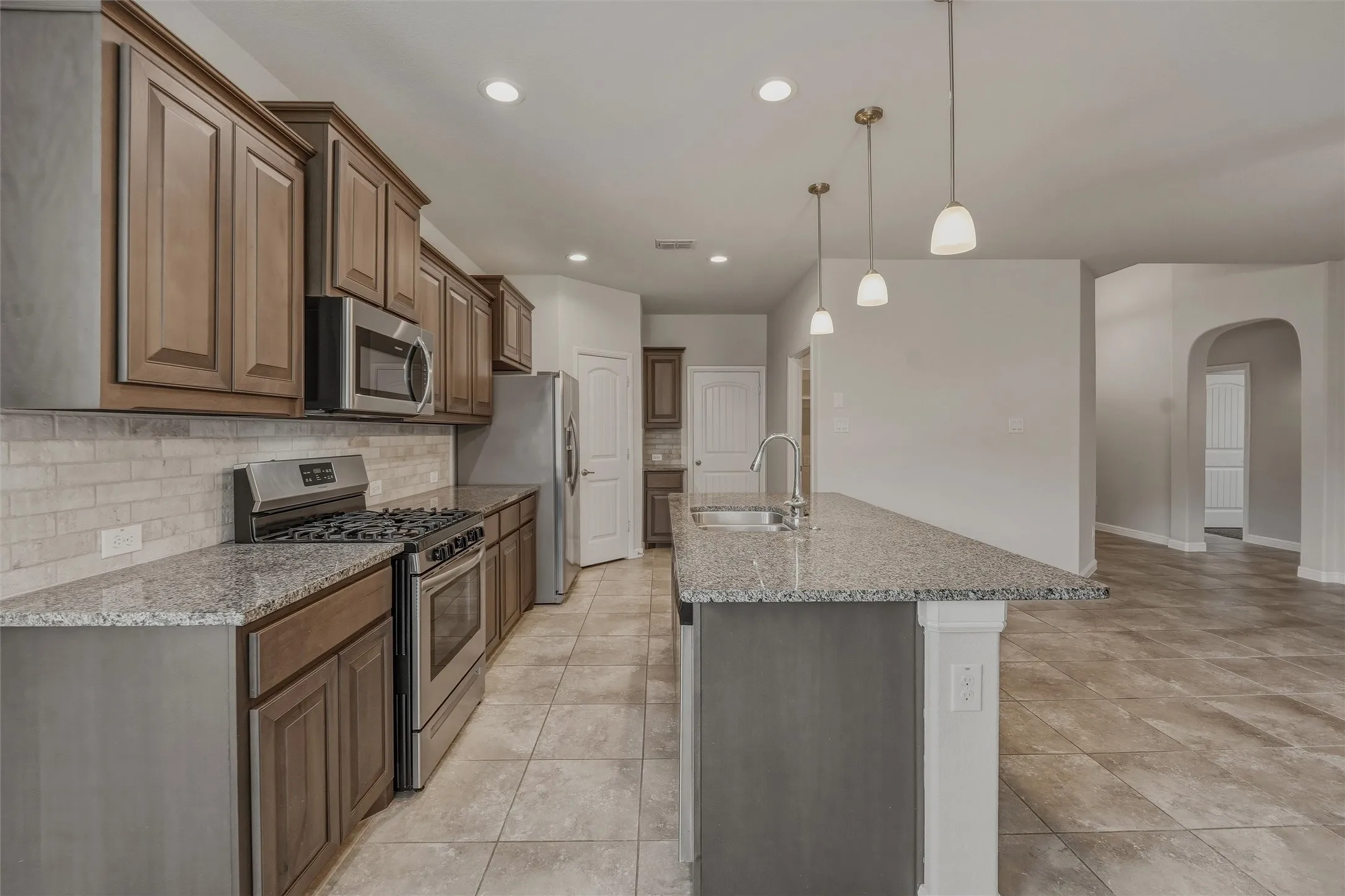 Open-concept kitchen featuring an expansive center island with a stainless steel sink, granite countertops, wood cabinetry, stainless steel appliances, and a tiled backsplash