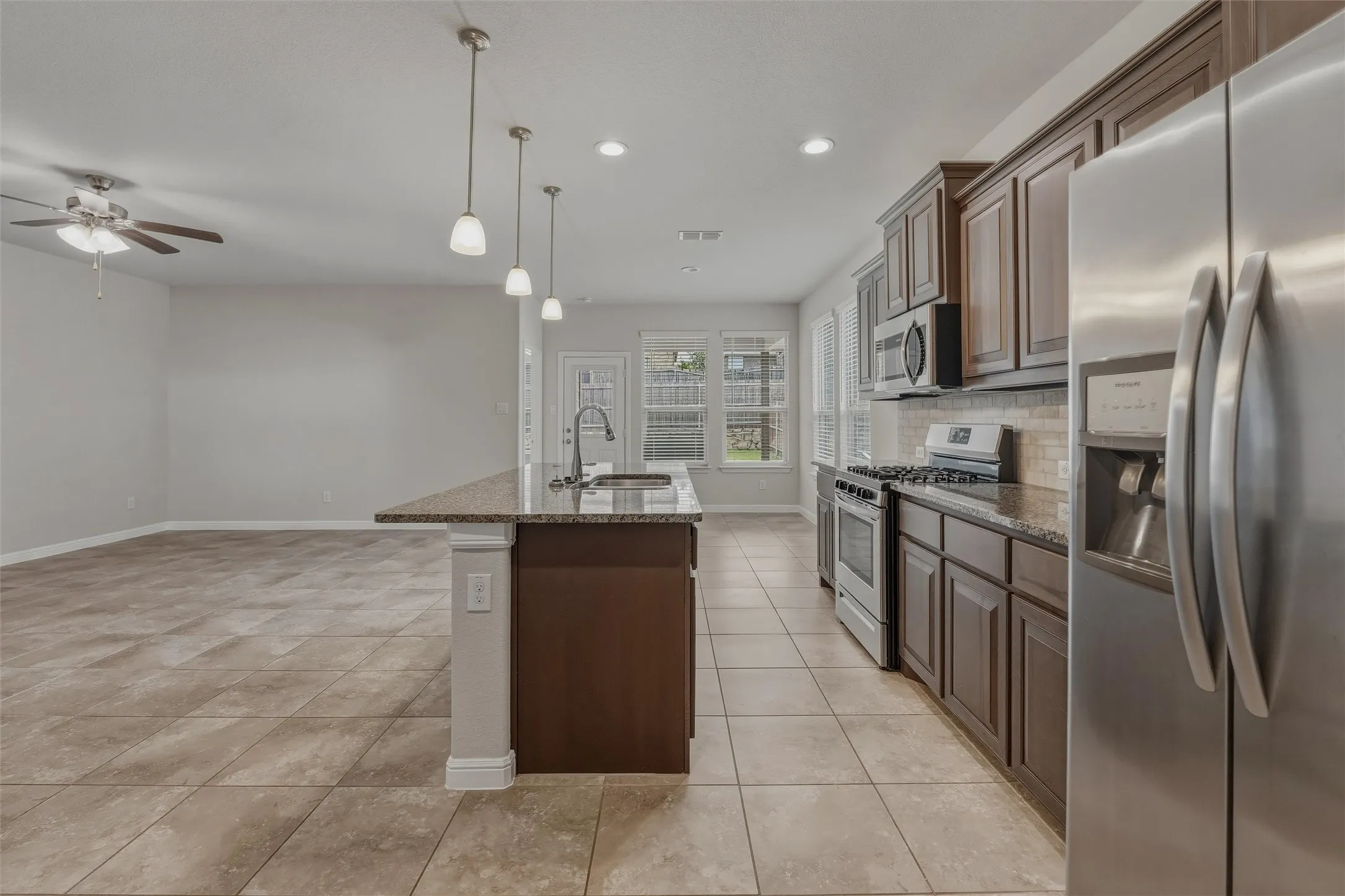 Open-concept kitchen featuring an island with a sink and pendant lighting, stainless steel appliances, dark wood-finish cabinetry, and a tile backsplash