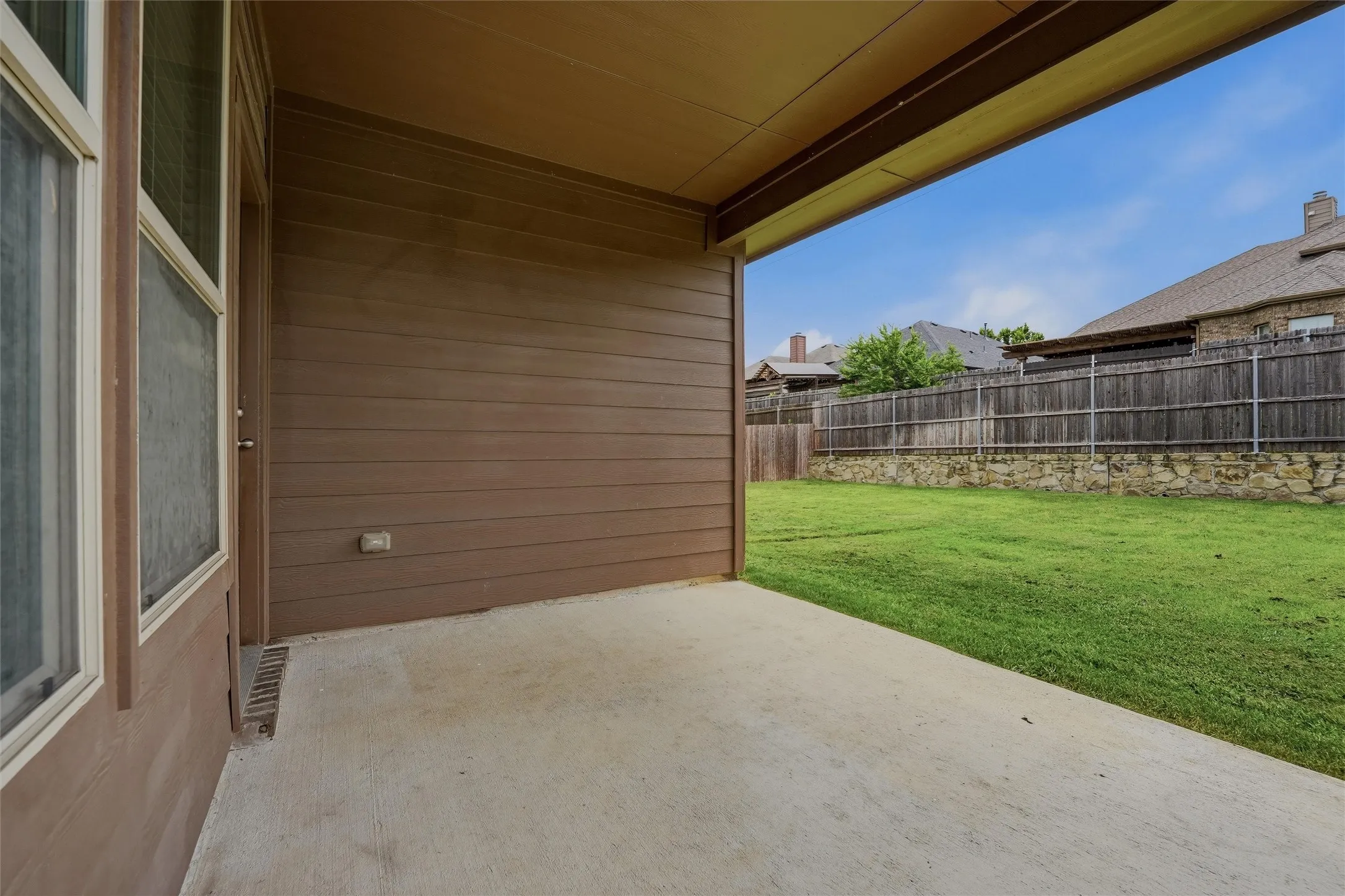 Covered concrete patio opening to a spacious lawn with a wood fence and stone retaining wall