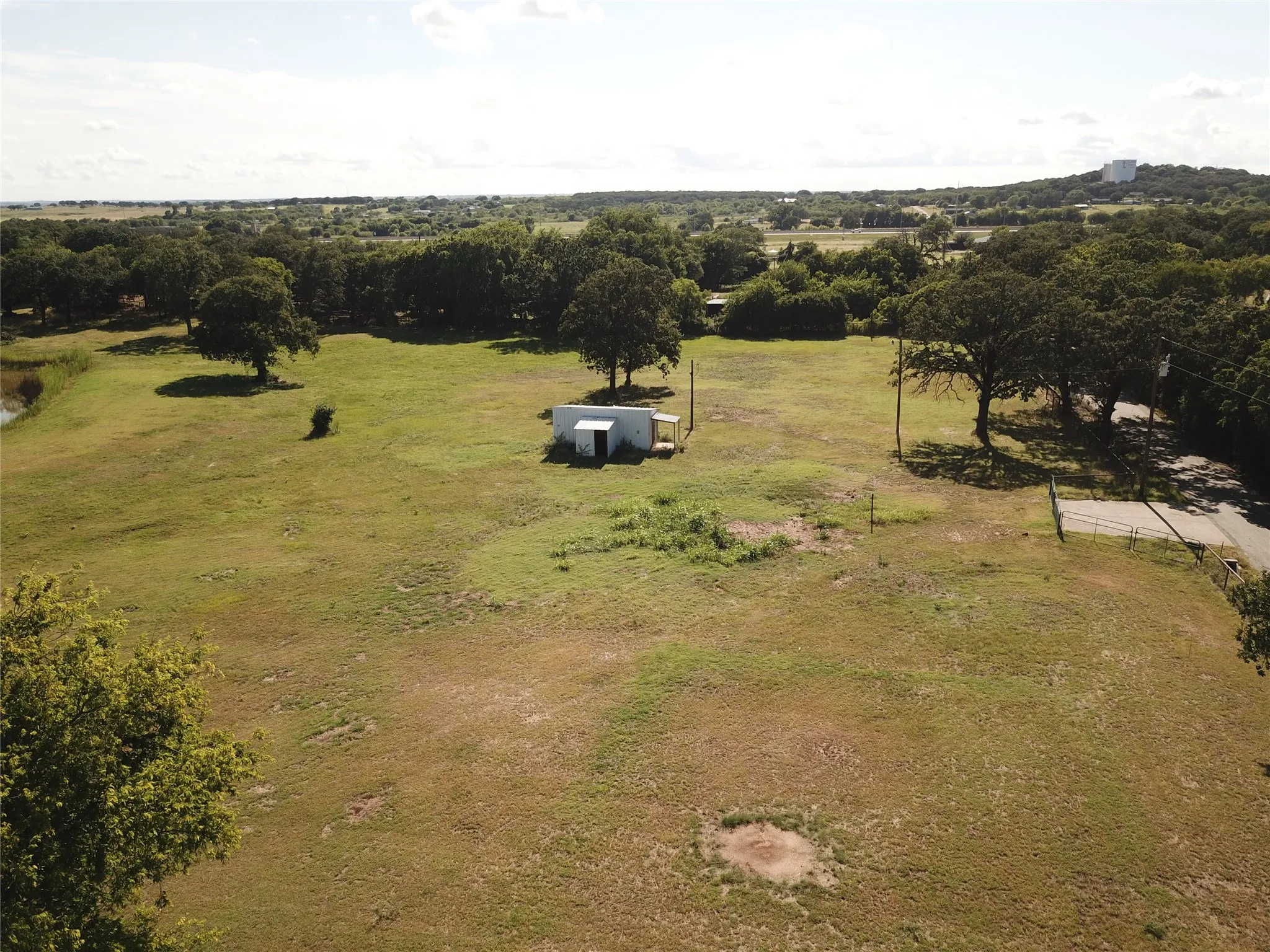 Expansive land parcel featuring open fields, mature trees, a small utility building, and road frontage