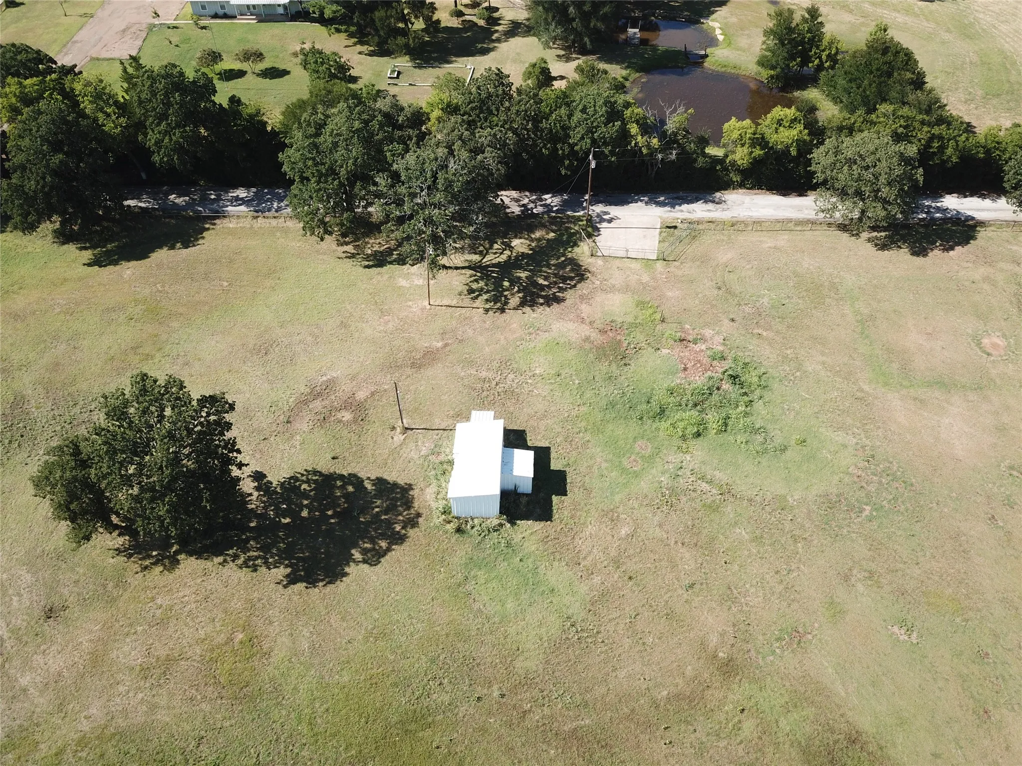 Expansive land parcel featuring native grassy areas, mature trees, a small outbuilding, and concrete approach to county road. Electrical transformer installed with interior power pole and light