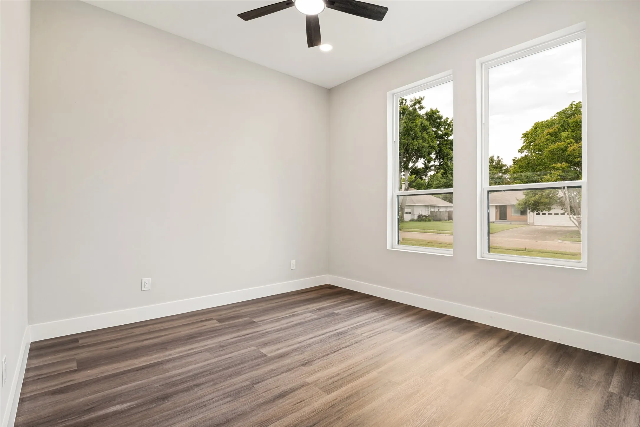 Room featuring wood-finish flooring, light neutral wall paint, two tall windows, a ceiling fan with integrated lighting, and white baseboards