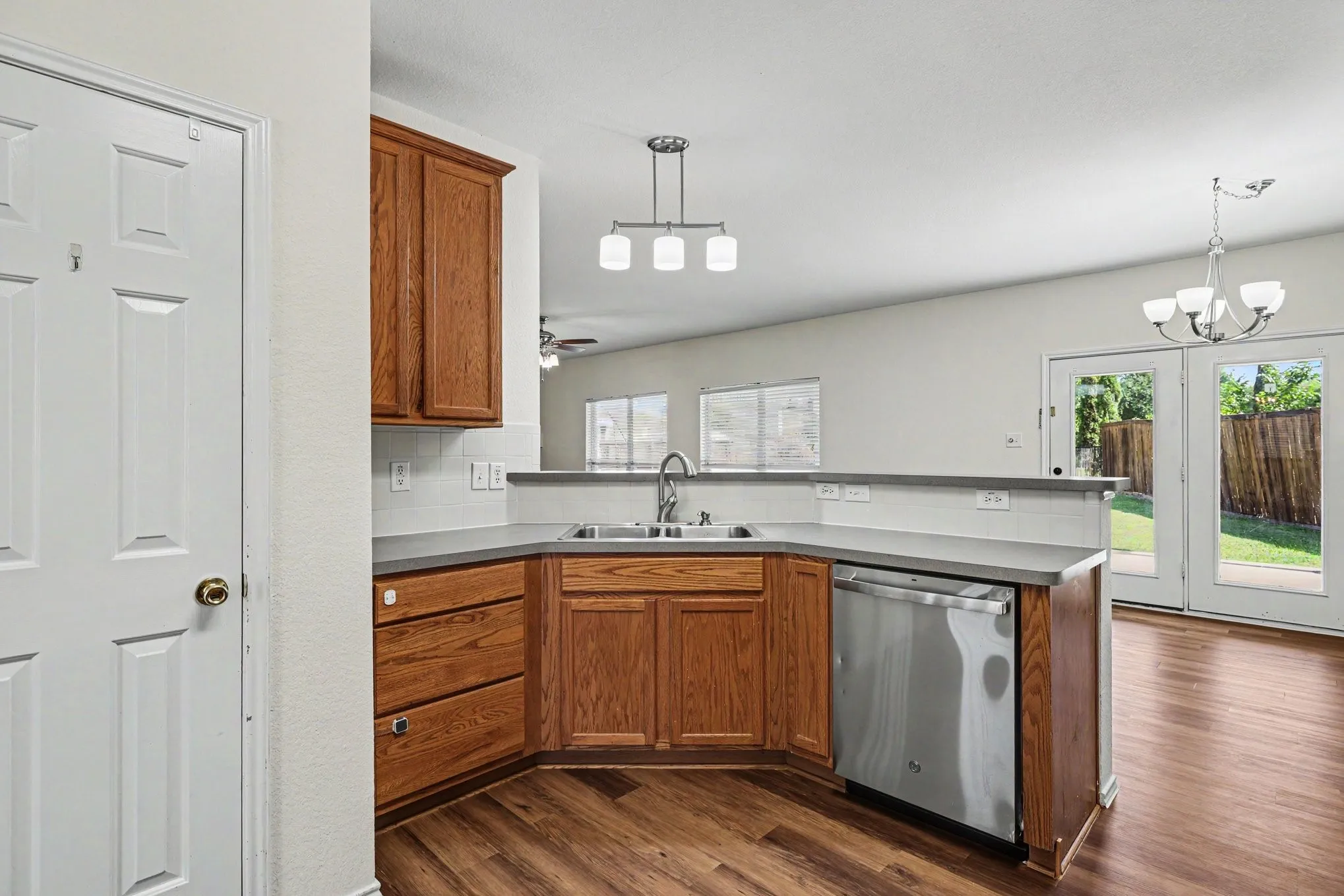 Kitchen featuring wood-finish flooring, oak cabinetry, a stainless steel dishwasher, a dual basin sink, and a peninsula with a light-toned countertop and backsplash