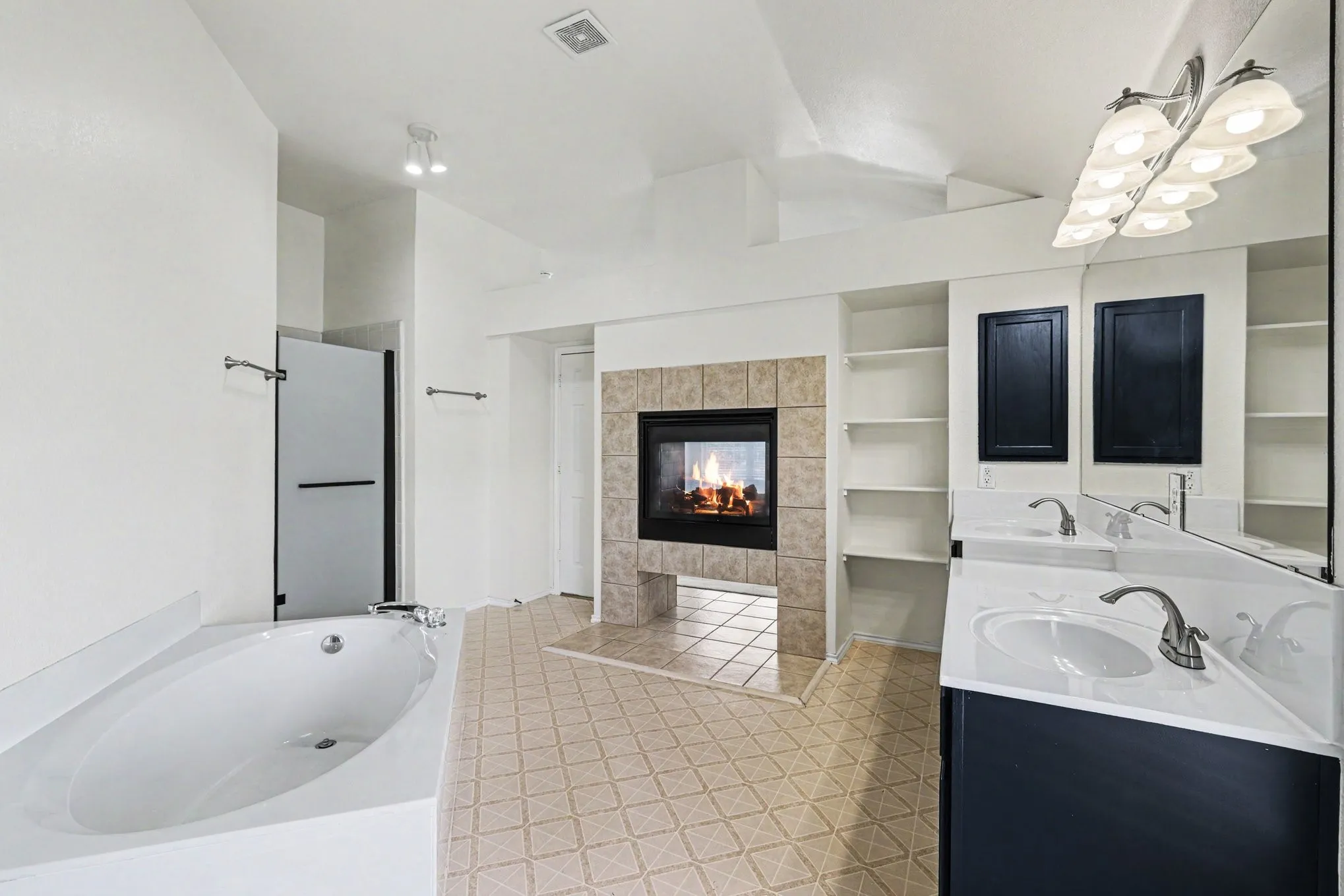 Spacious bathroom featuring a built-in soaking tub, a tiled fireplace with a mantel, a dual vanity with white countertops, integrated shelving, and patterned tile flooring