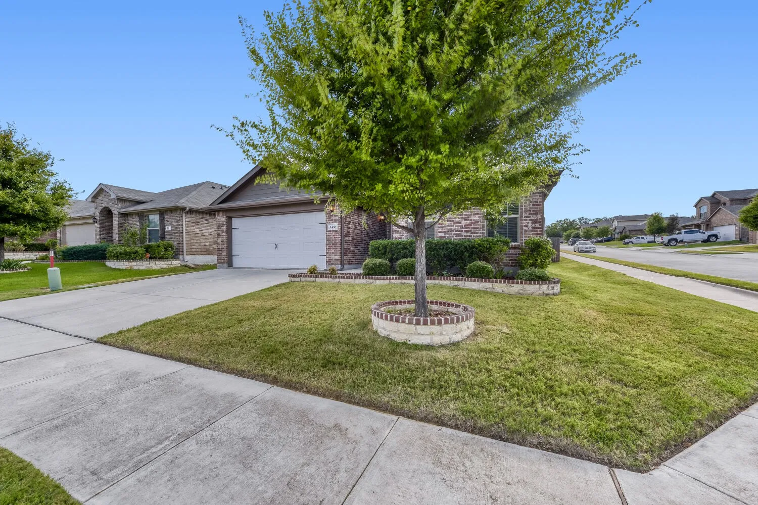 Brick facade residence featuring an attached two-car garage, concrete driveway, and front yard landscaping with mature trees