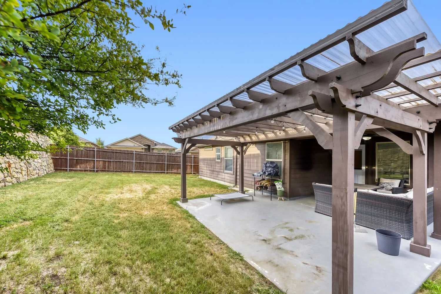 Private backyard featuring a concrete patio with a wood pergola, clear corrugated roof, and a stone retaining wall