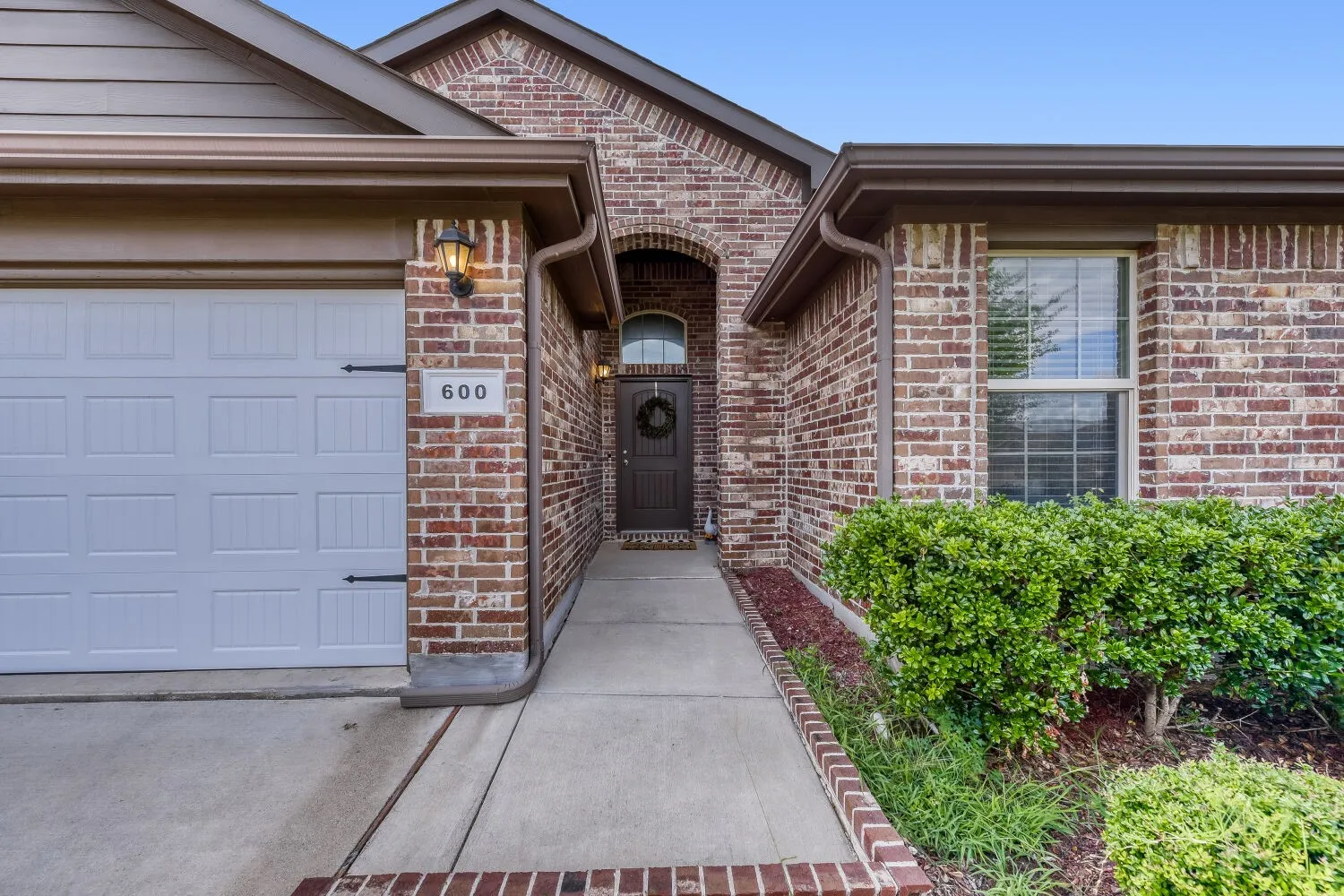 Brick exterior featuring a paneled garage door, archway entry, front door with an arched transom window, and established perimeter shrubs