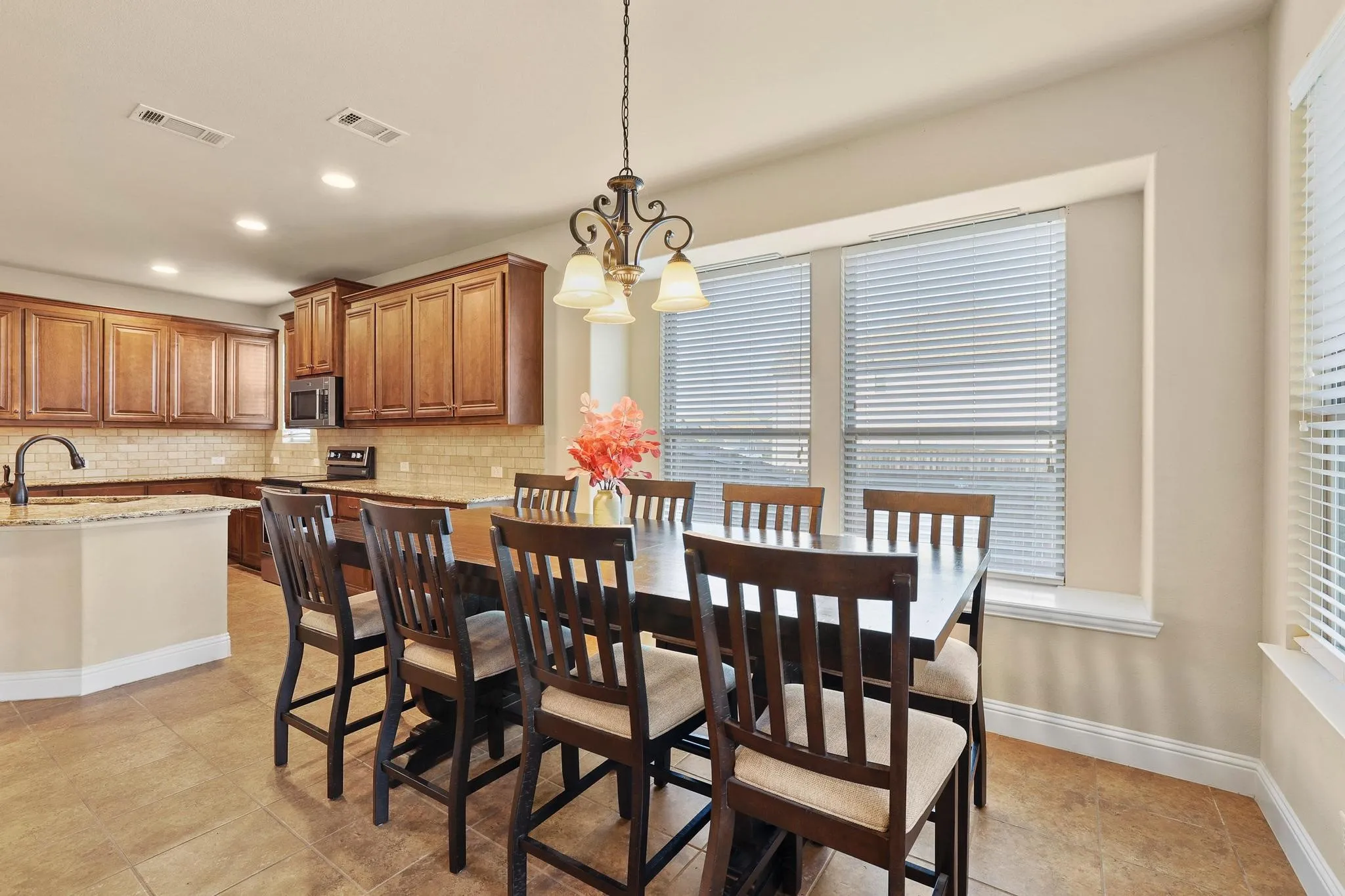 Open-concept dining area featuring a chandelier and multiple windows, complementing the adjacent kitchen with wood cabinetry, light-toned backsplash, granite countertops, and built-in microwave