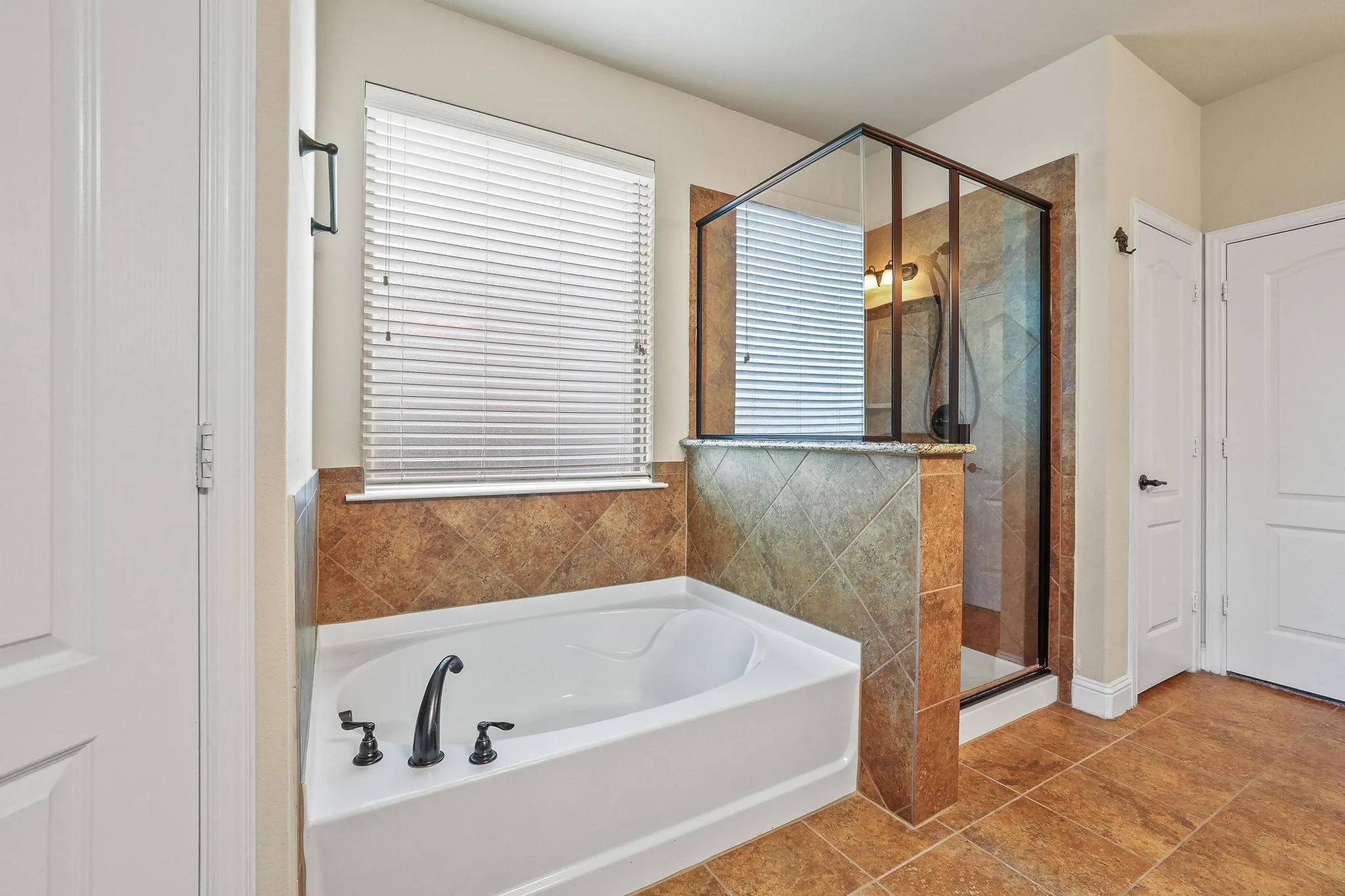 Bathroom featuring a white soaking tub with dark-finish fixtures, a separate glass-enclosed shower, and earth-toned tile flooring and surrounds