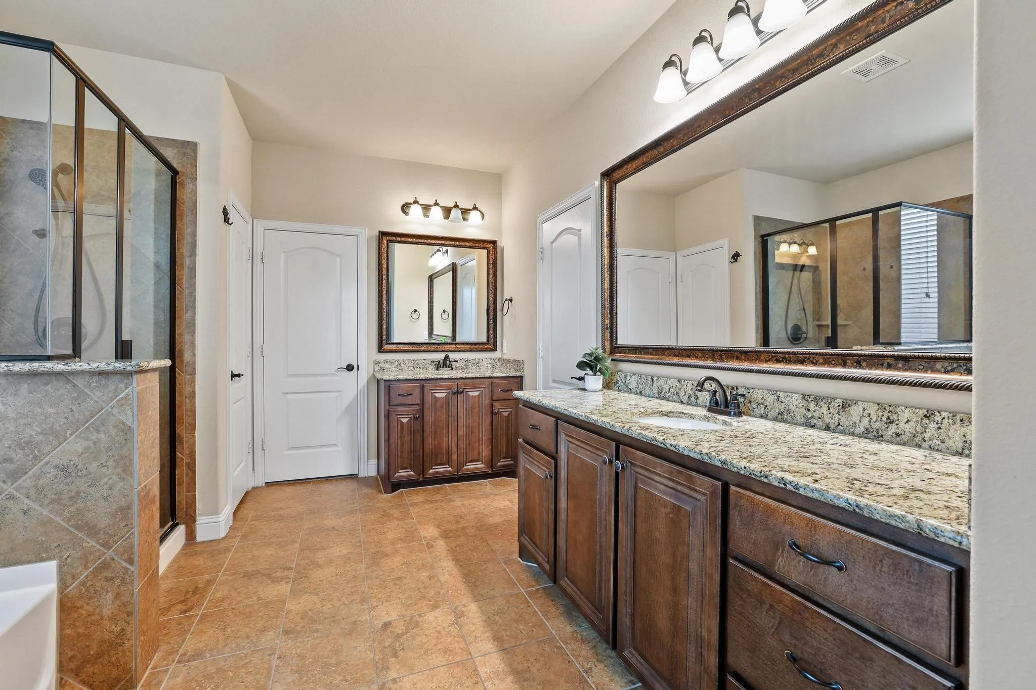 Spacious bathroom featuring dual vanities with granite countertops, rich wood cabinetry, tile flooring, and a glass-enclosed shower with tiled walls