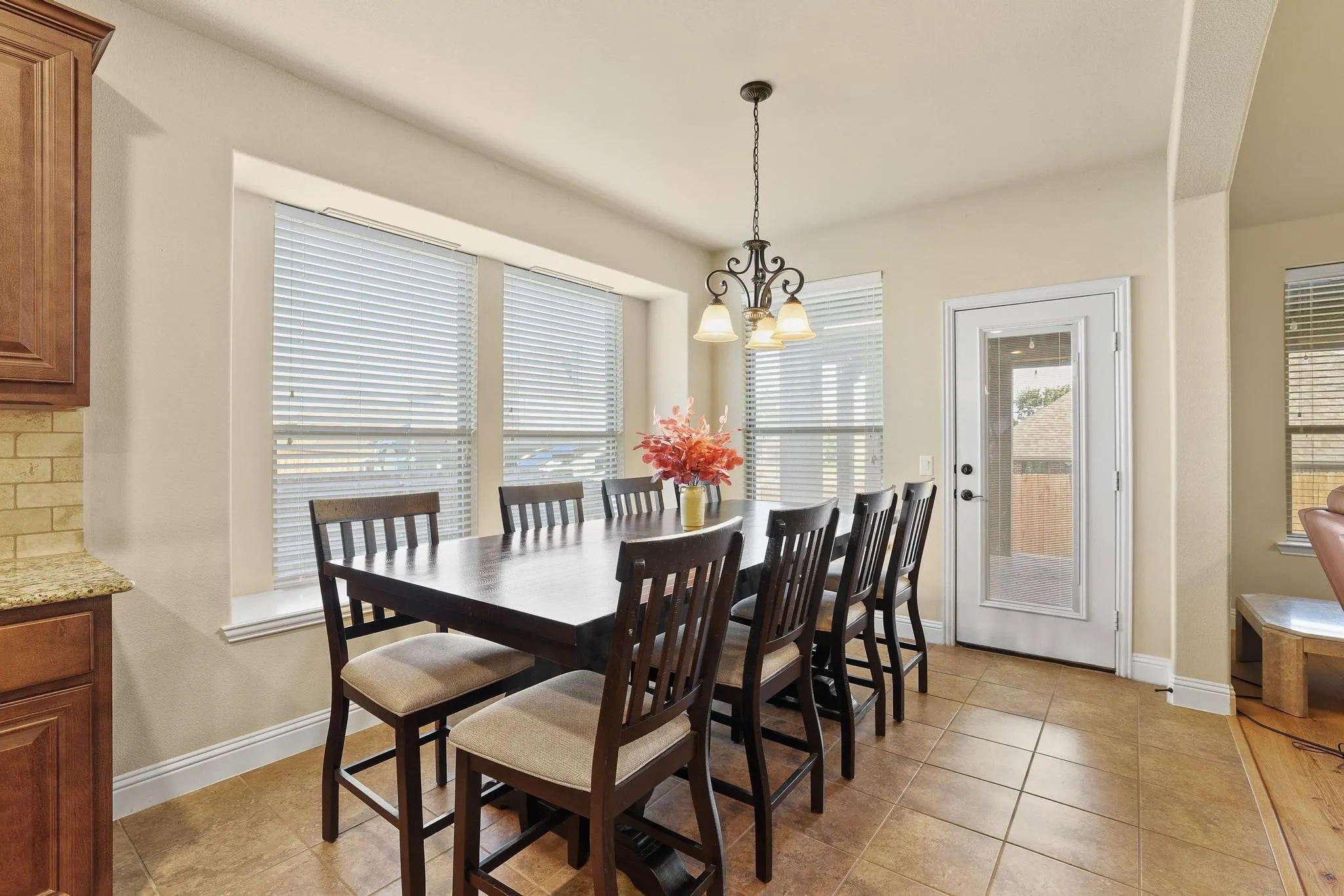 Bright dining area featuring a pendant light fixture, large windows with blinds, a glass-paneled exterior door, tile flooring, and a portion of a kitchen with wood cabinetry and a tile backsplash