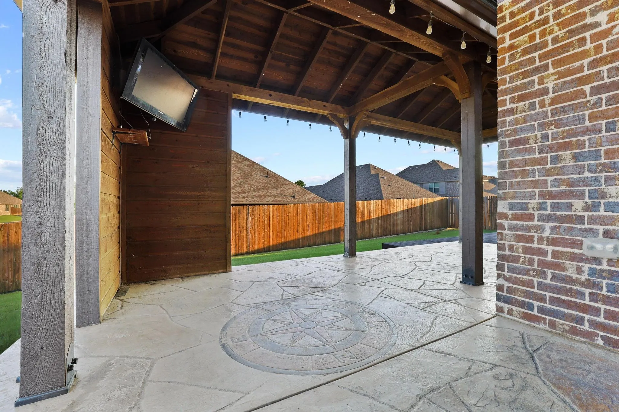 Covered patio featuring stamped concrete flooring with a compass rose inlay, string lighting, exposed wood ceiling beams, and a mounted flat-screen display