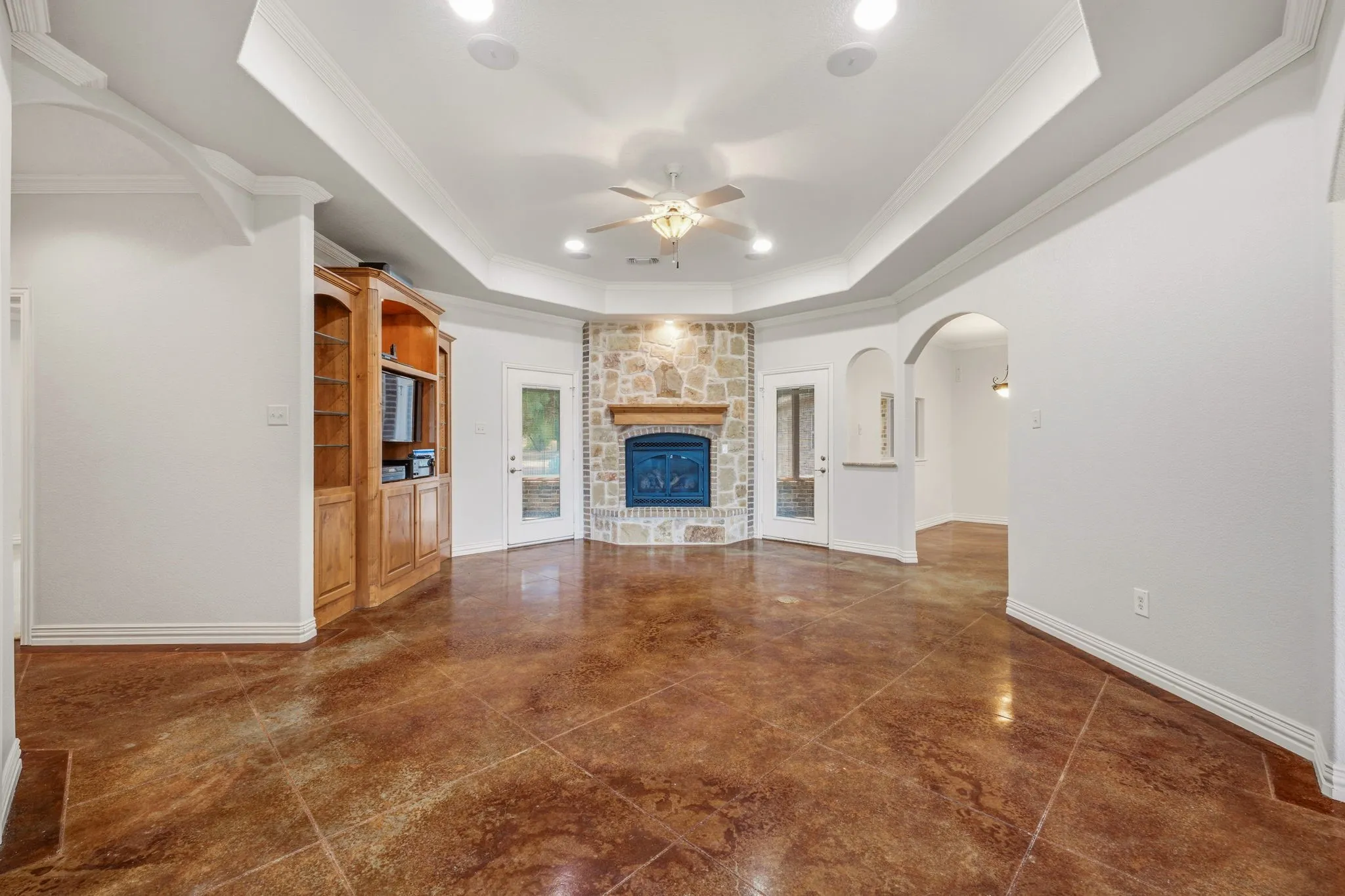 Spacious room featuring a stone fireplace with a wood mantel, stained concrete flooring, a tray ceiling with recessed lighting, and two glass-paneled doors