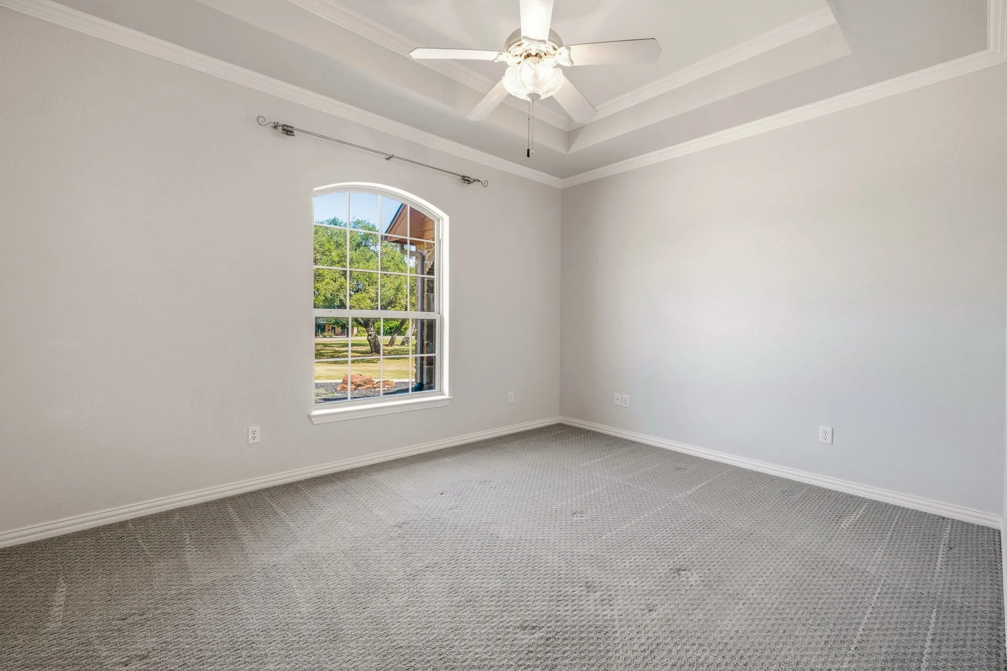 Spacious room featuring a tray ceiling with crown molding, an arched window, and a ceiling fan with integrated lighting