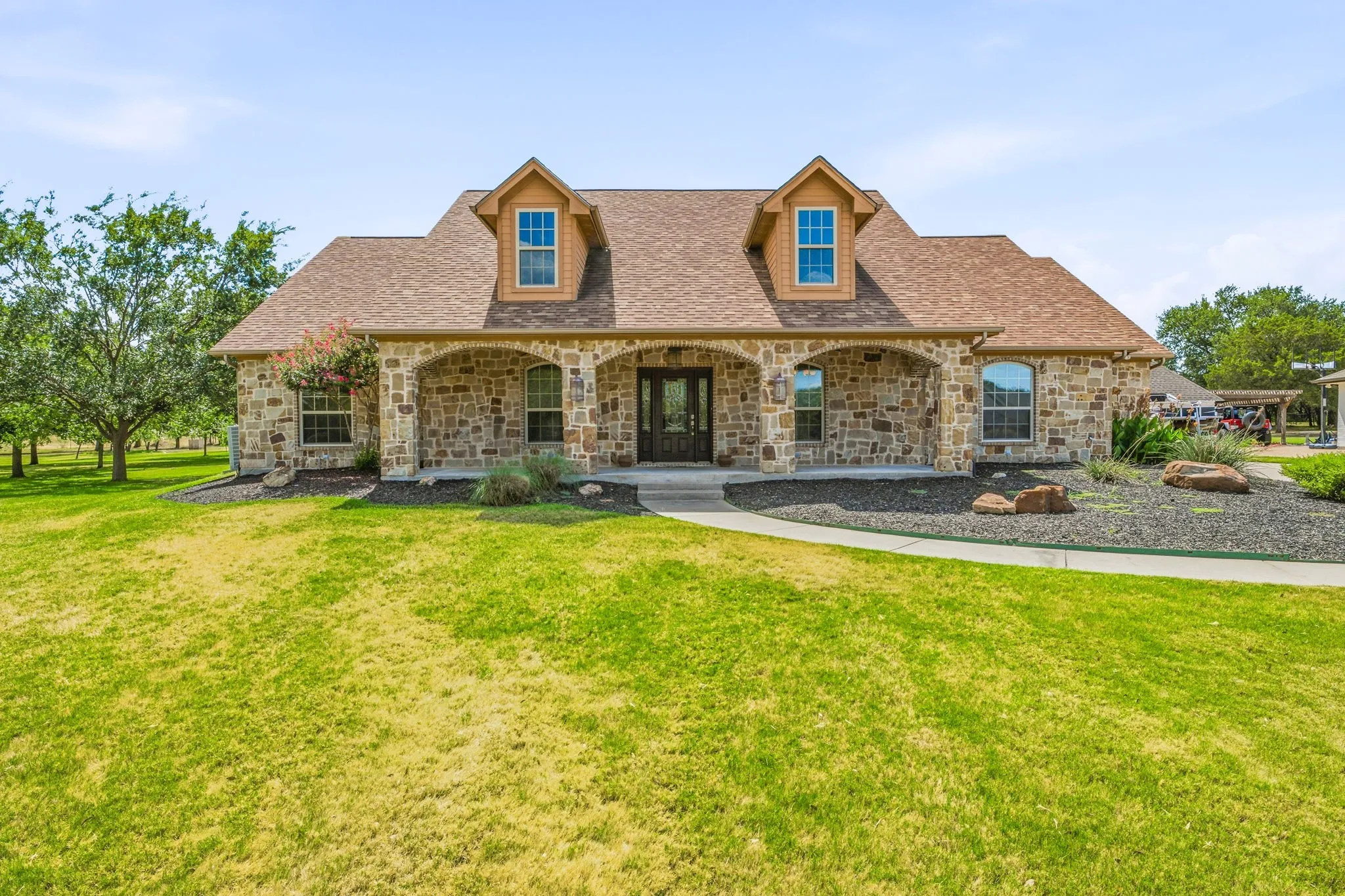 Stone facade residence featuring a covered front porch, dormer windows, a shingle roof, and a concrete pathway