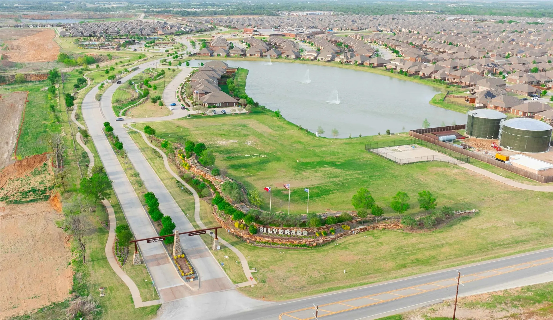 Aerial perspective of a planned community featuring a central lake with water features, a prominent entry monument with landscaping, and curvilinear roadways