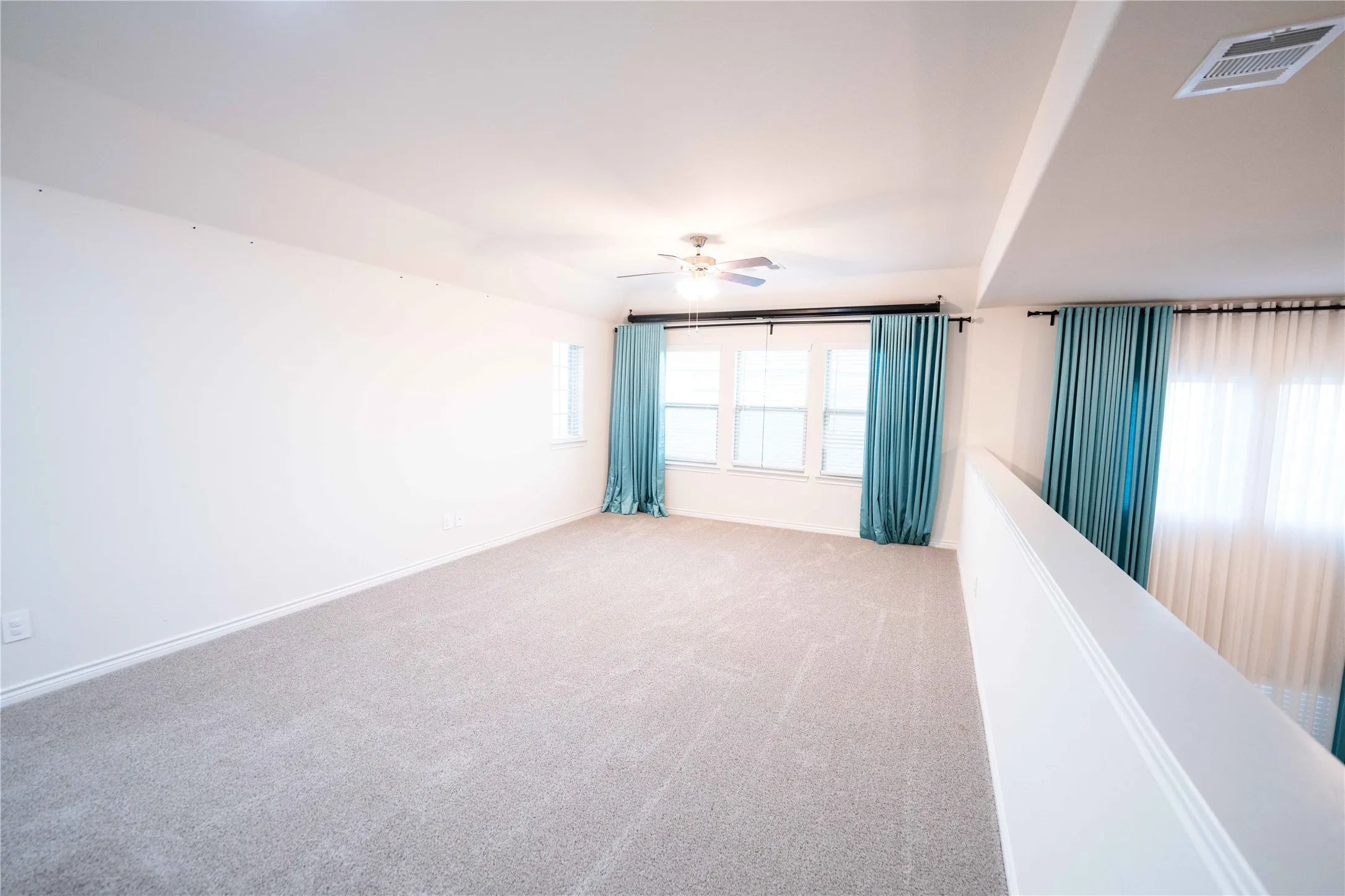 Spacious upper-level room featuring light gray carpeting, multiple windows with teal drapery, a ceiling fan, and white painted walls