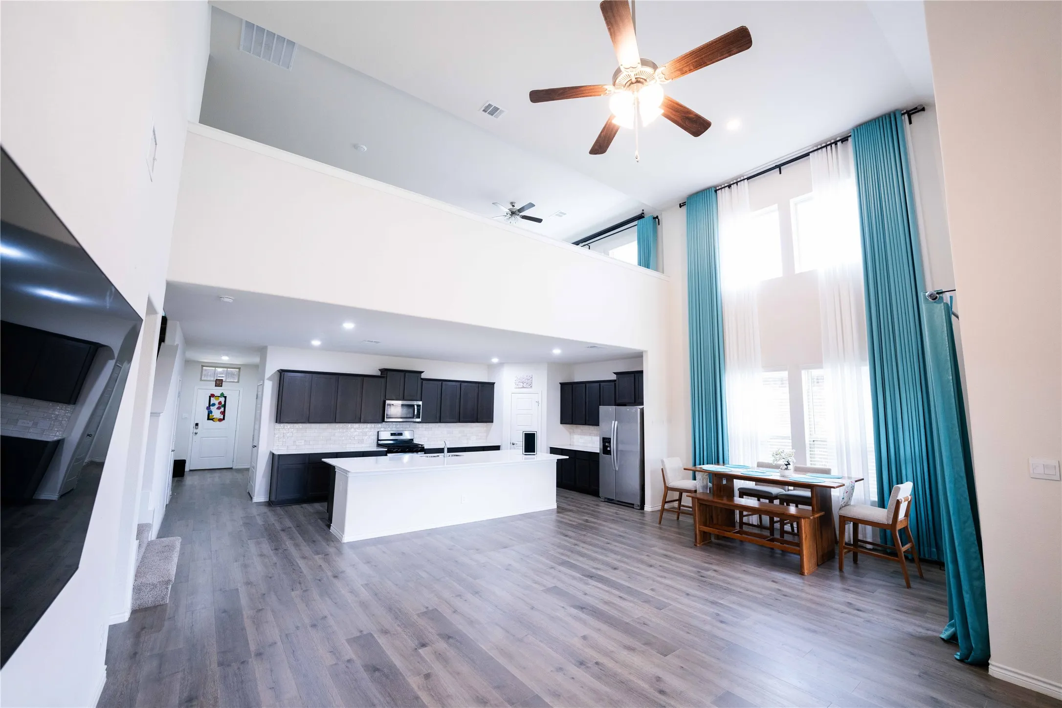 Open-concept living space featuring high ceilings, wood-finish flooring, a contemporary kitchen with dark cabinetry and a white island, and tall windows with bright curtains